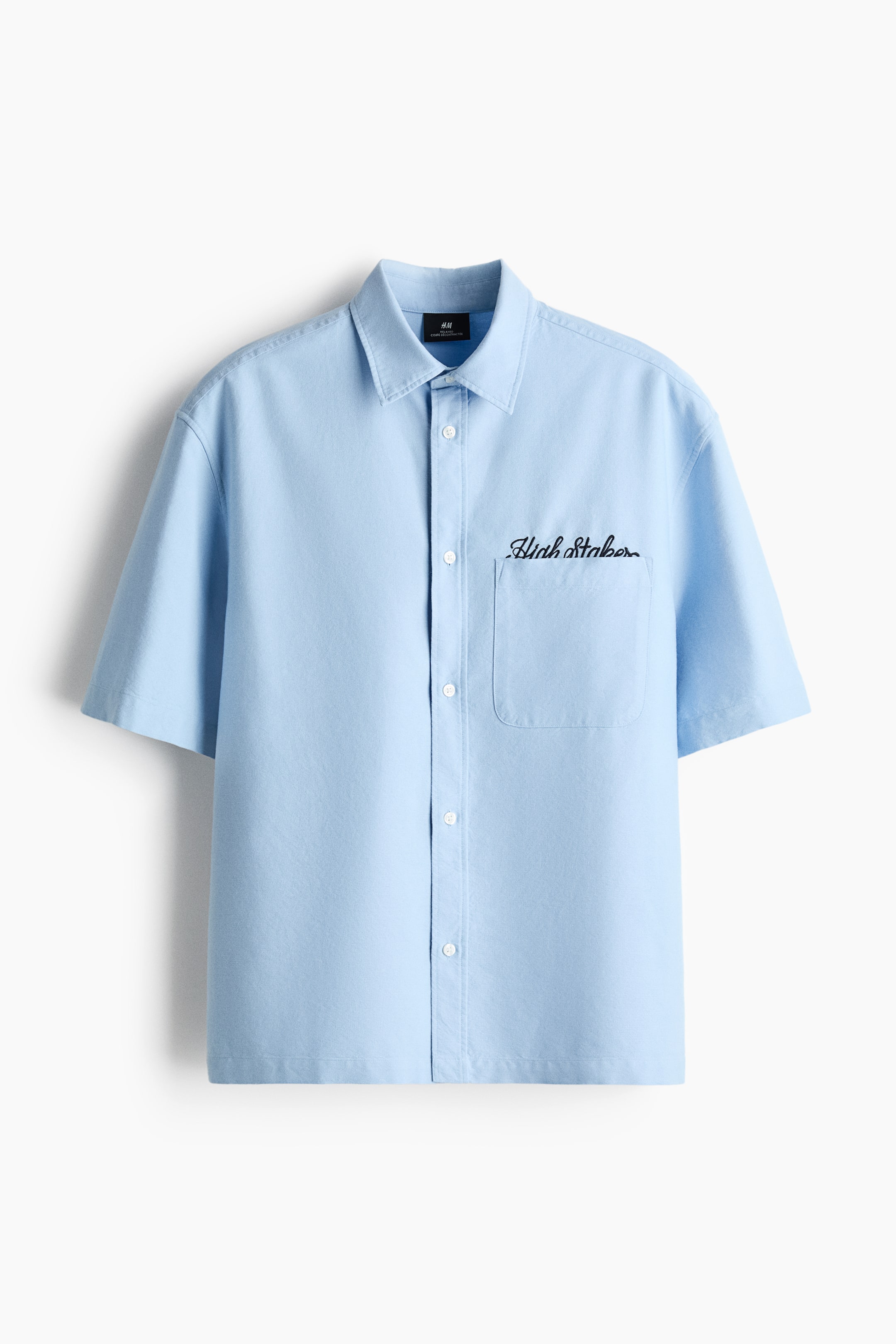 View larger image: A light blue short-sleeved shirt is shown from the front, featuring a pointed collar, short sleeves, and a full button-front closure with white buttons. A chest pocket appears on the left side, with black script embroidery reading "High Staker" above it.