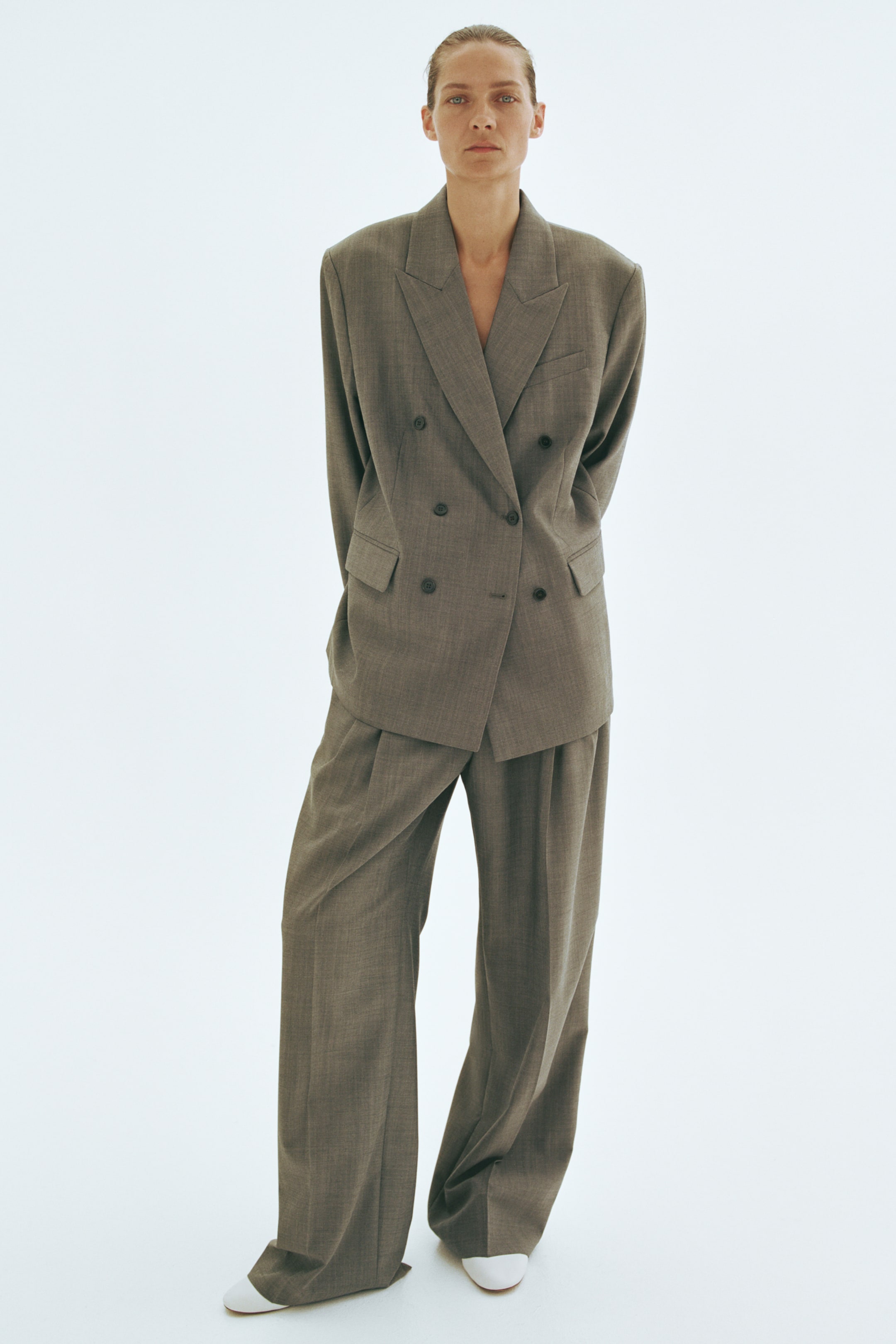 Visualizza immagine più grande: I pantaloni in tessuto misto lana grigio-marrone indossati dalla modella presentano una linea dritta e ampia, con pieghe sul davanti e pieghe nette. I pantaloni sono abbinati a un completo coordinato con blazer doppiopetto e scarpe bianche.