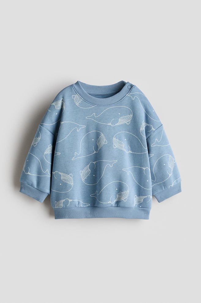 Un sweatshirt albastru imprimat prezintă PARTEA FRONTALĂ, având un model pe întreaga suprafață format din contururi albe de balenă. Este prevăzut cu guler rotund reiat, două capse pe umărul stâng, manșete reiate și tiv reiat. - 1