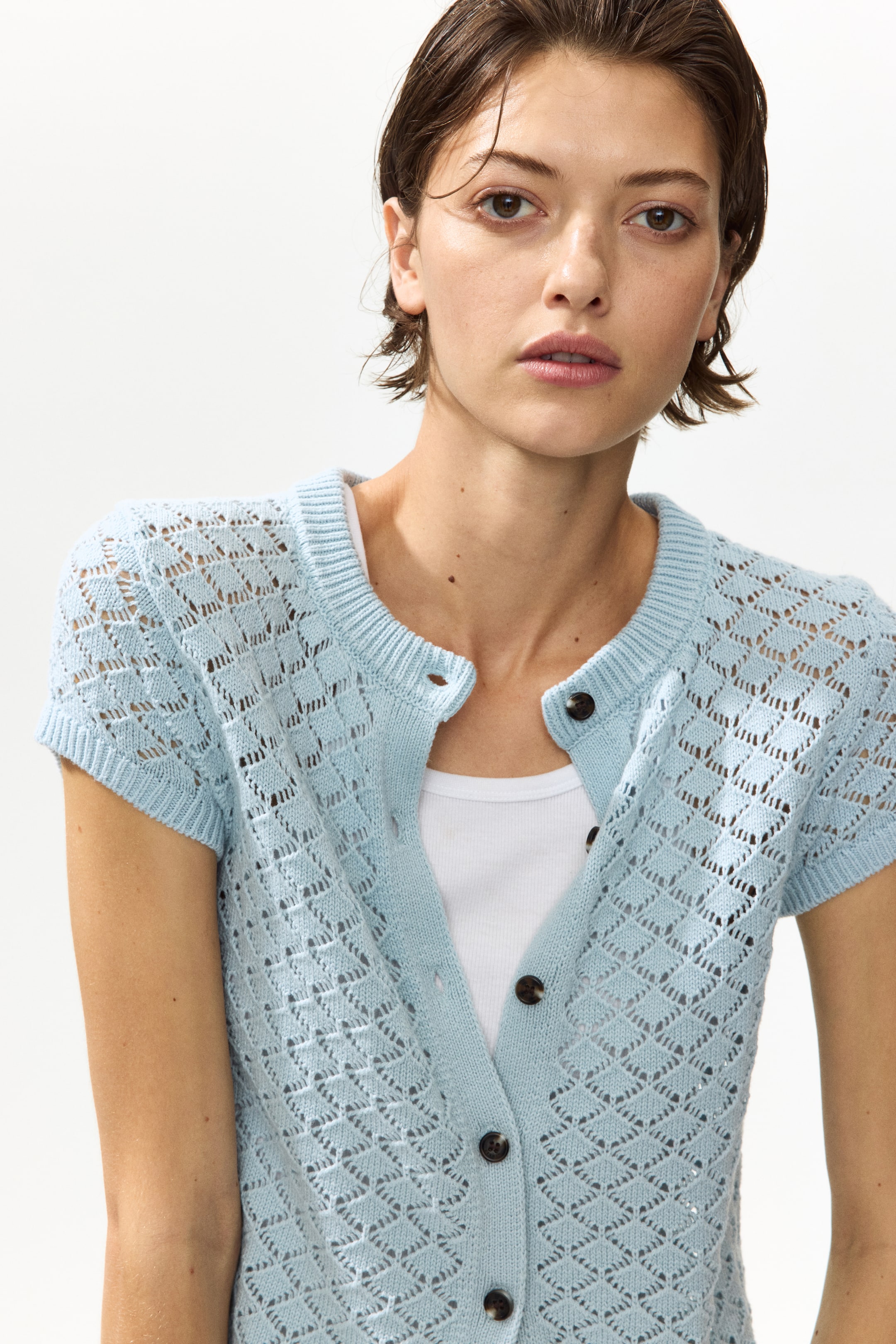 Größeres Bild anzeigen: Ein hellblauer, kurzer, figurbetonter Pointelle-Strick-Cardigan wird vom Model getragen und über einem weißen Tanktop geschichtet. Der Cardigan verfügt über einen runden, gerippten Ausschnitt, Kappärmel mit gerippten Bündchen und dunkle Knöpfe an der Vorderseite, von denen einige derzeit offen sind.