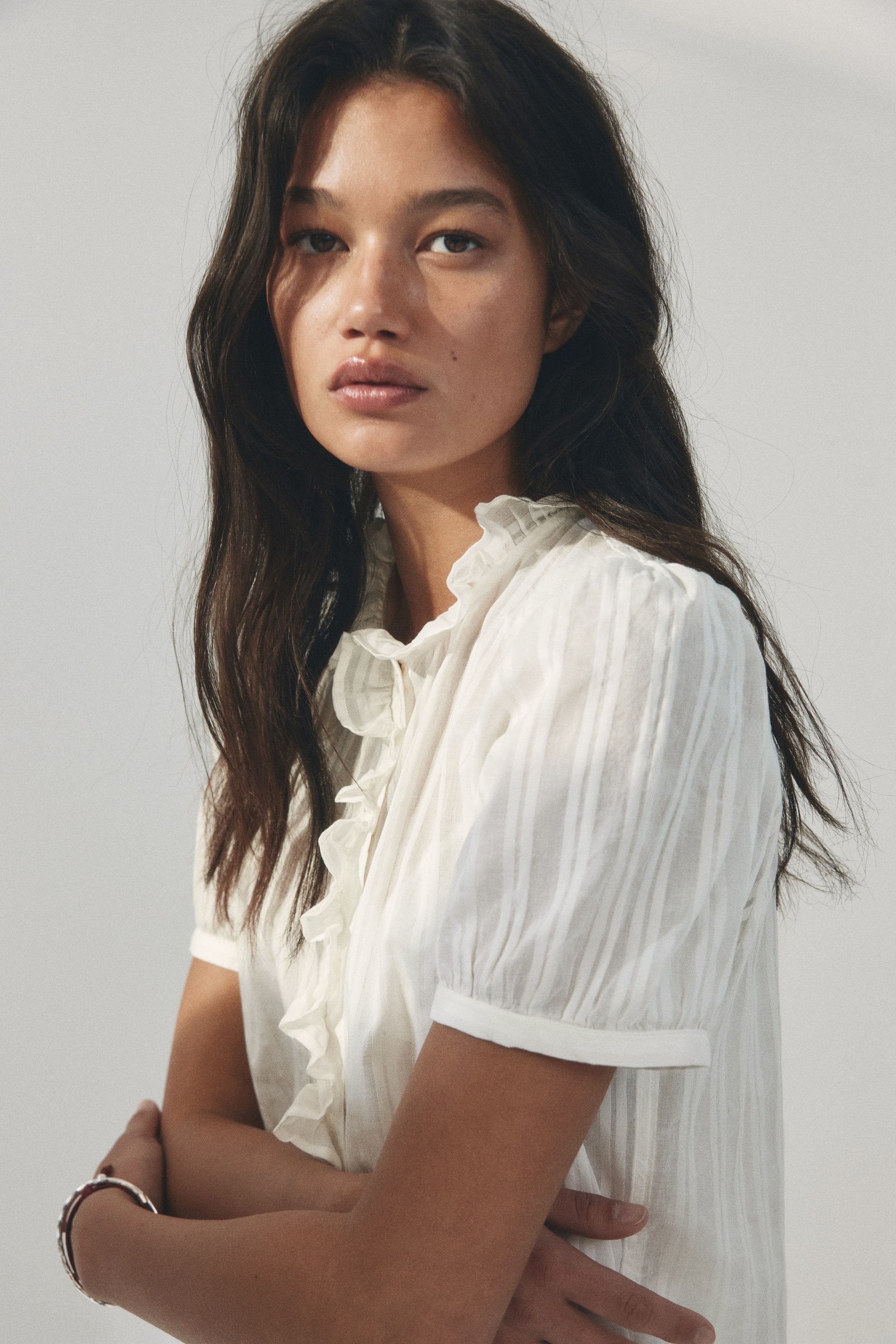 View larger image: A white blouse featuring a high ruffled collar with a tie detail and a ruffled placket down the front. It is designed with short, puffed sleeves and a subtle vertical striped texture, displayed from a slightly front-facing angle.