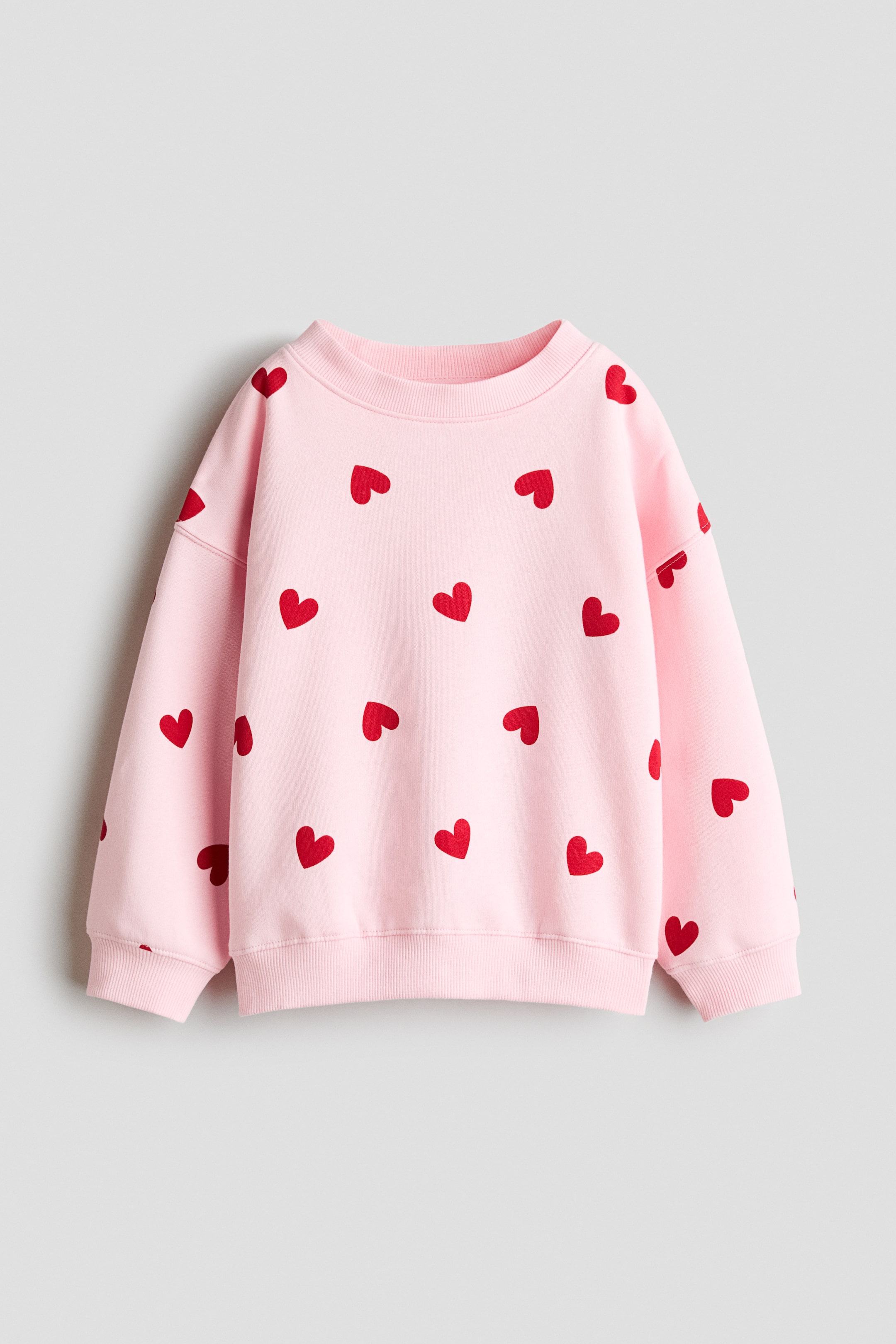View larger image: A pink sweatshirt showcases a pattern of red hearts across its front, featuring a round, rib-trimmed neckline. It is crafted with dropped shoulders, long sleeves, and ribbing at the cuffs and hem.