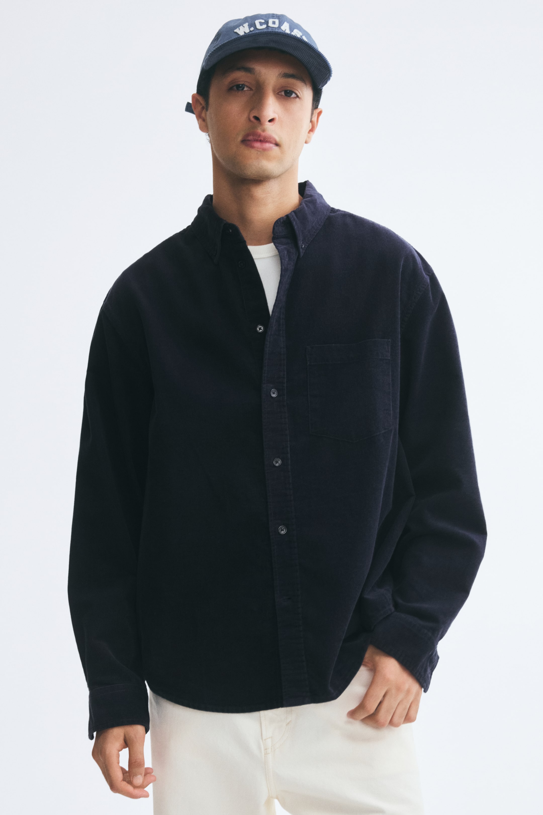 View larger image: A dark navy-blue corduroy shirt is being worn by the model over a white crewneck T-shirt and light-colored trousers. The shirt features a button-down collar left slightly open at the neck, a button-front closure, and a single open chest pocket.