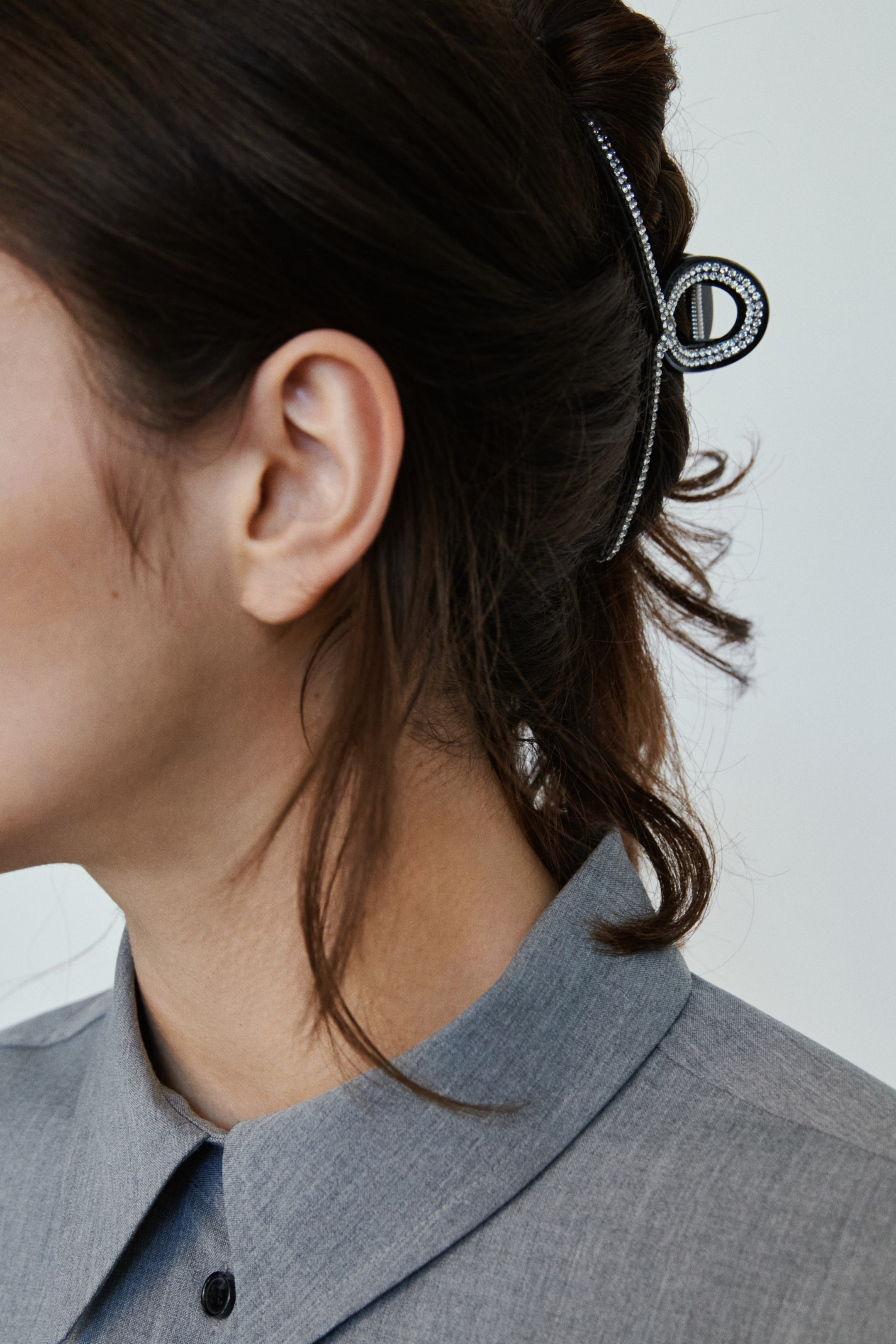 View larger image: A black plastic hair claw with rhinestone embellishments is securing the model's dark brown hair, which is styled in an updo, with some loose strands falling around the neck.