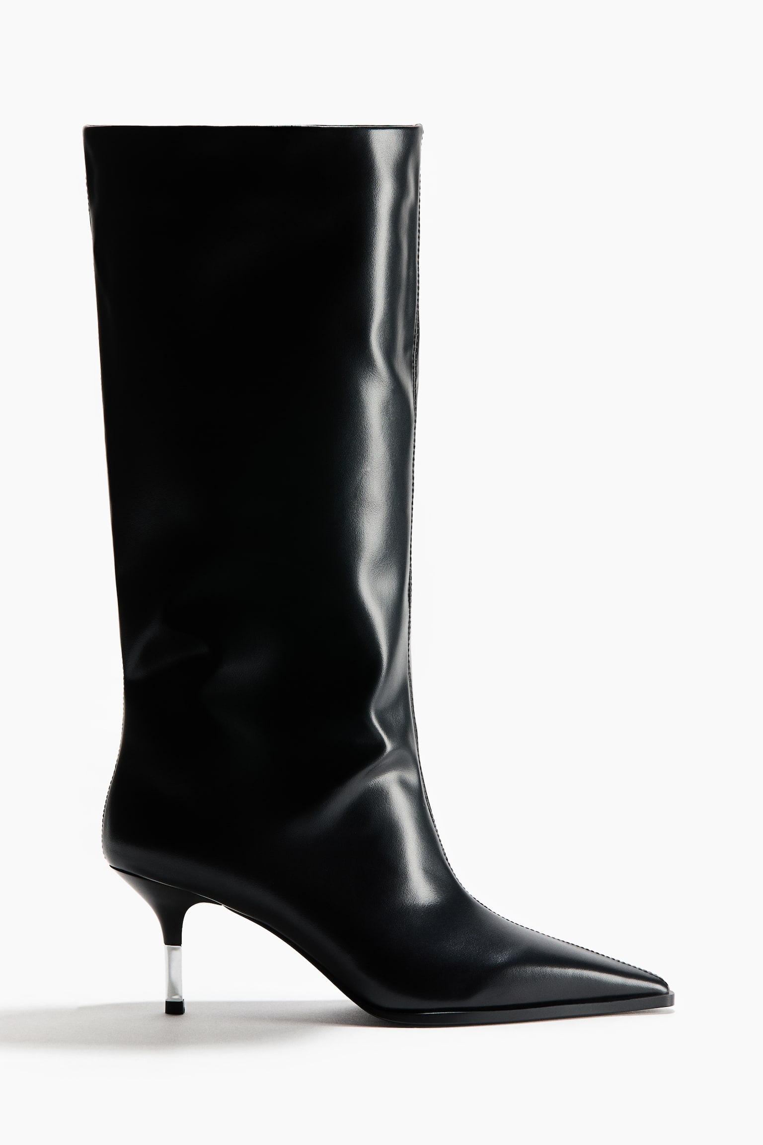 Una bota negra hasta la rodilla aparece de perfil lateral, mostrando una puntera afilada, una costura central y una caÃ±a ancha y estructurada. La bota presenta un tacÃ³n bajo tipo kitten heel con una parte superior transparente y translÃºcida.
