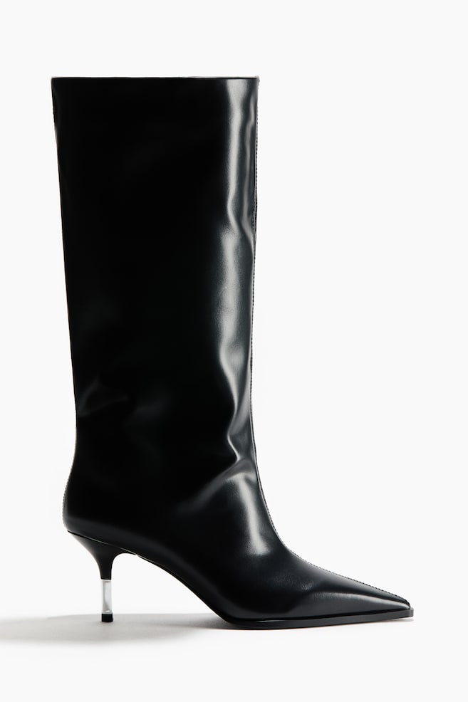 A black knee-high boot is shown in side profile, featuring a pointed toe, a central seam and a wide, structured shaft. The boot has a low kitten heel with a clear, transparent upper section. - 1