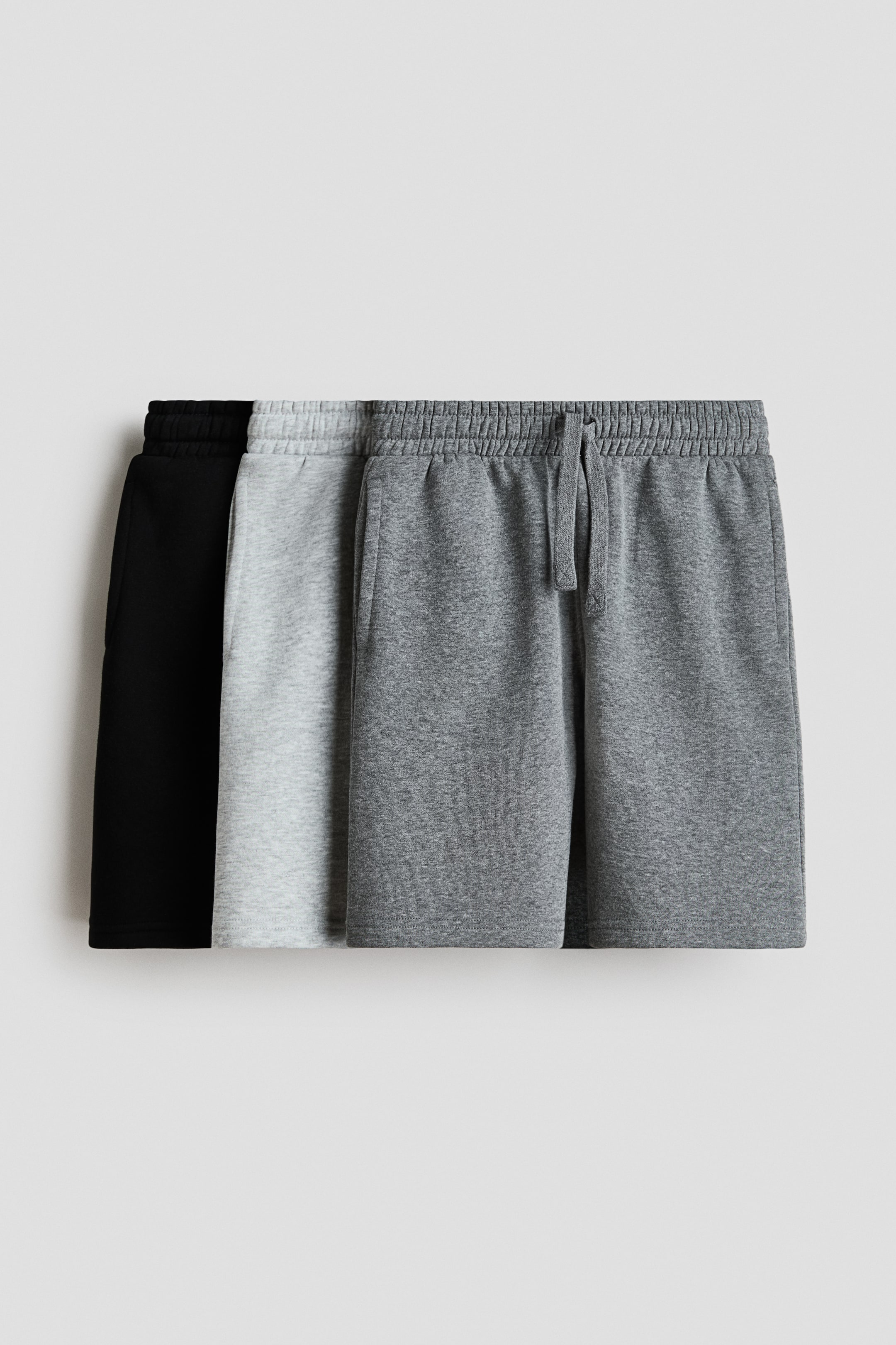 View larger image: Three pairs of shorts are showcased, crafted from lightweight sweatshirt fabric. The front-facing dark grey pair features an elasticated, drawstring waist with a tie, and discreet side pockets, while a light grey and a black pair are partially visible behind it.