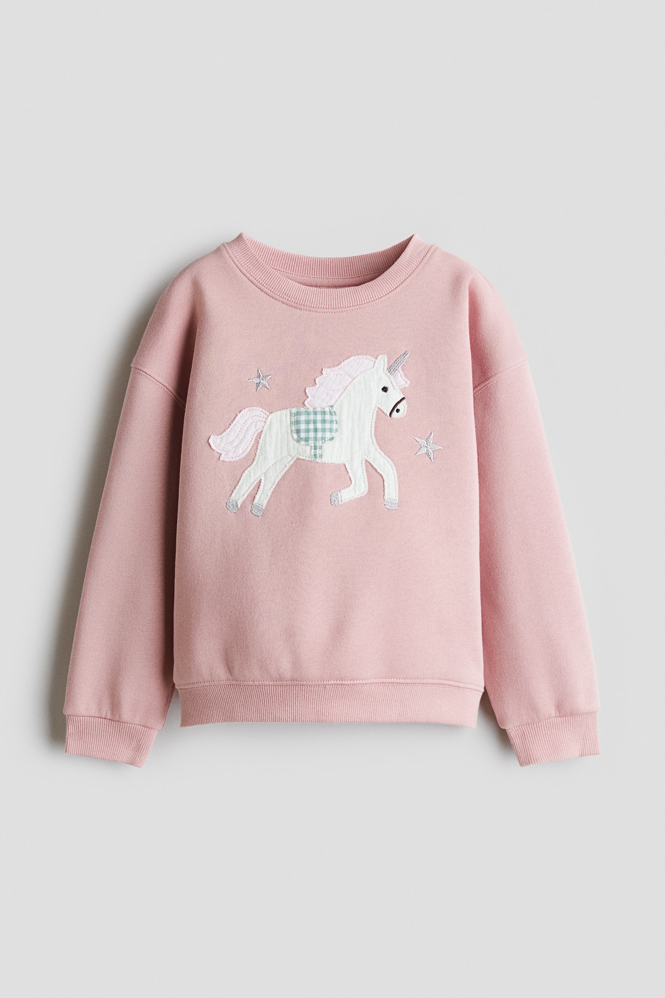 View larger image: A pink sweatshirt is shown from the front, featuring a round, rib-trimmed neckline, dropped shoulders, ribbed cuffs, and a ribbed hem. A white unicorn motif with a light green checkered saddle and light pink mane and tail is centred on the front, flanked by two small grey stars.