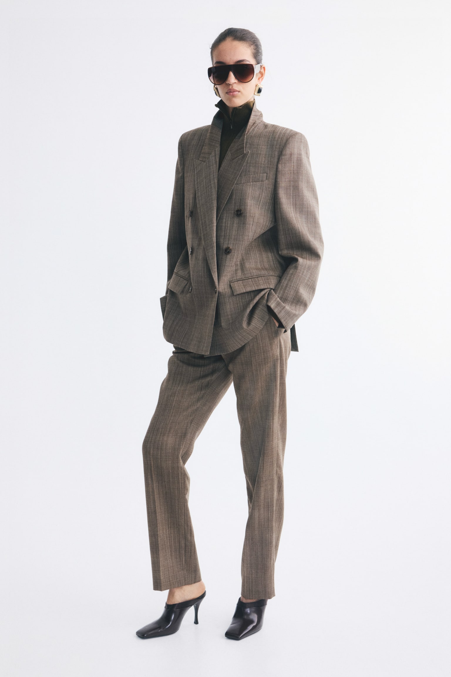 Brown wool-weave trousers with slightly tapered legs and front creases are worn by the model. The trousers are styled with a matching double-breasted blazer and black heeled mules, with the model placing one hand in the pocket. - 1