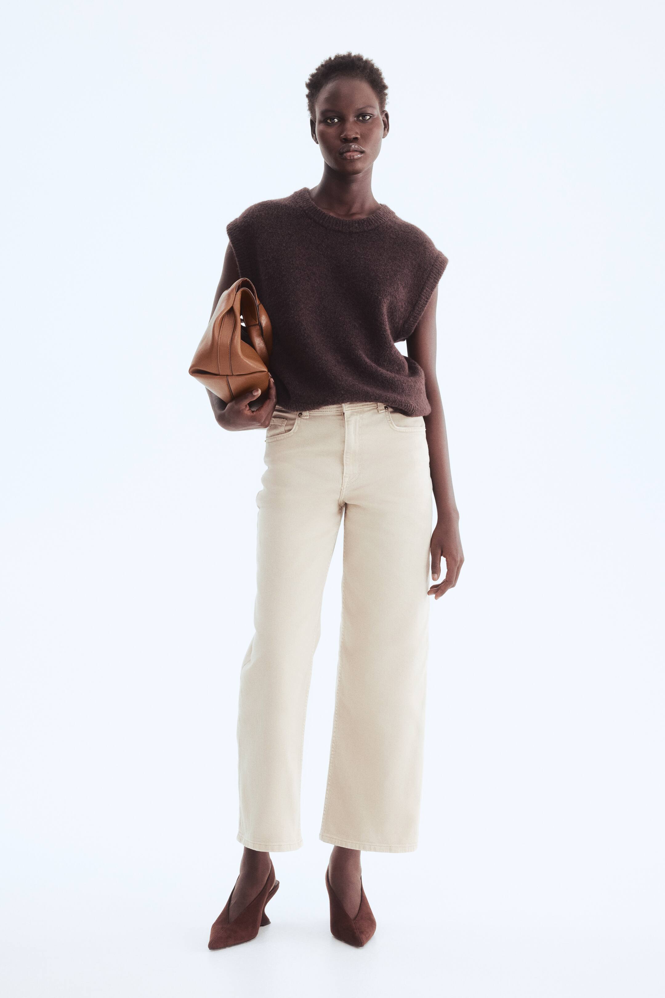 Agrandir l'image: Un gilet sans manches en maille pelucheuse marron foncé avec un col rond est associé à un jean beige clair taille haute à jambes larges. Des escarpins slingback en daim marron foncé à bout pointu avec des talons sculpturaux sont portés, et un sac à main en cuir beige avec une poignée supérieure pliée est tenu.