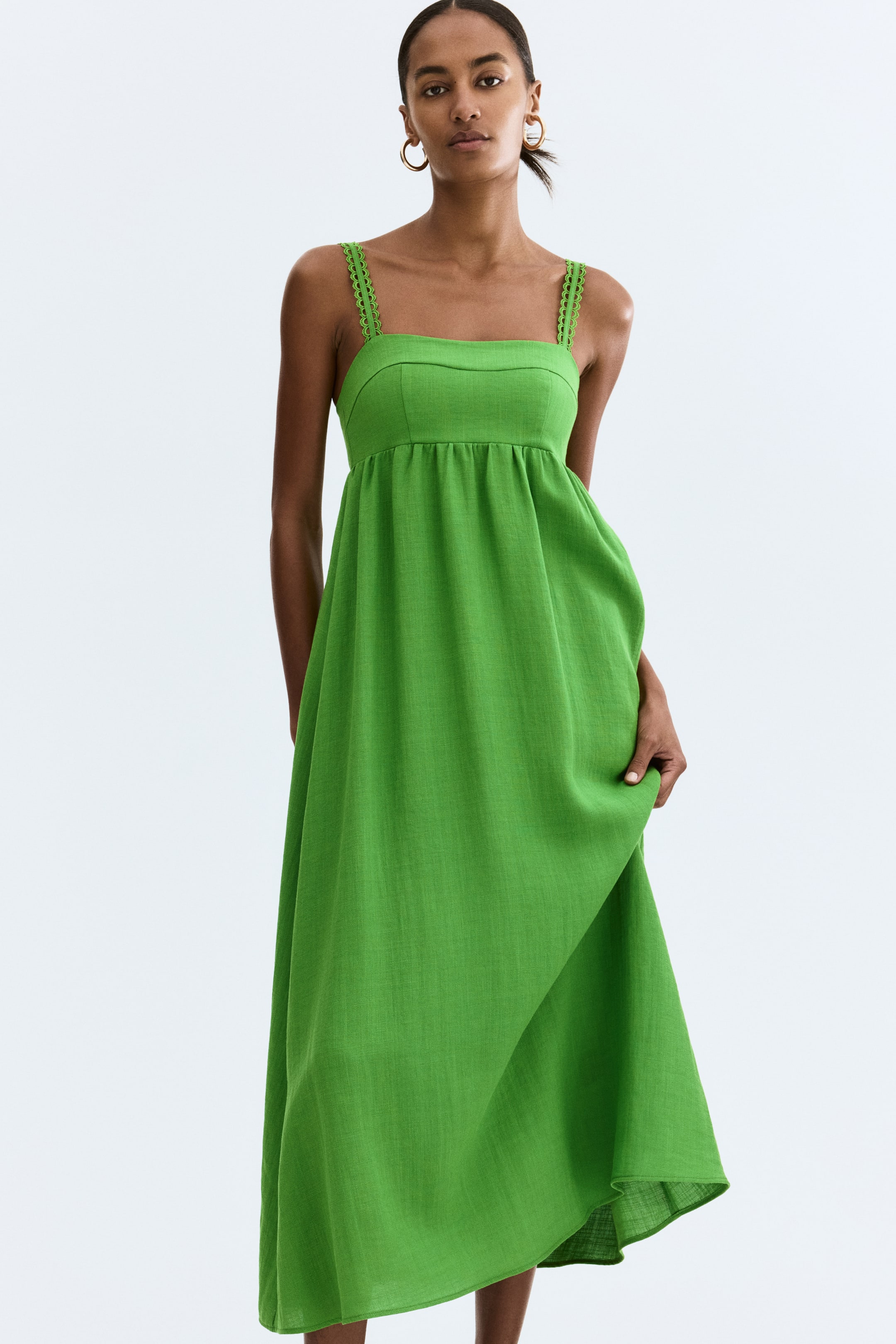 Ampliar la imagen: Un vestido midi verde brillante presenta tirantes con bordes festoneados y un escote cuadrado, con un corpiño ajustado que fluye hacia una cintura imperio fruncida y una falda completa. Una mano levanta suavemente una parte de la falda, mostrando su caída y textura.