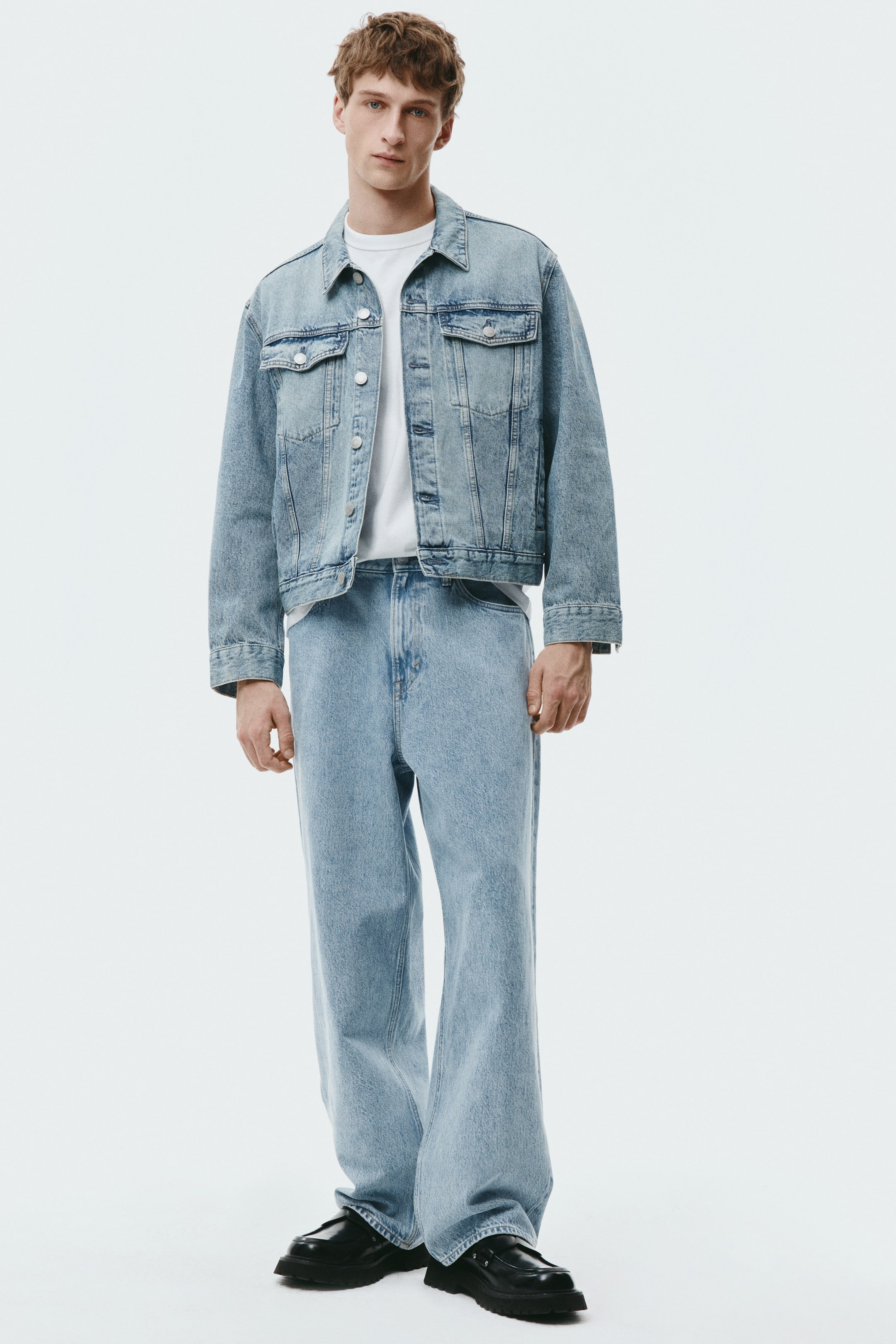 View larger image: The model wears light-blue 5-pocket jeans in rigid cotton denim, paired with a white T‑shirt and a matching denim jacket. The jeans feature a regular waist, a dropped crotch and a baggy fit with rounded legs that provide ample room and stack at the ankle.