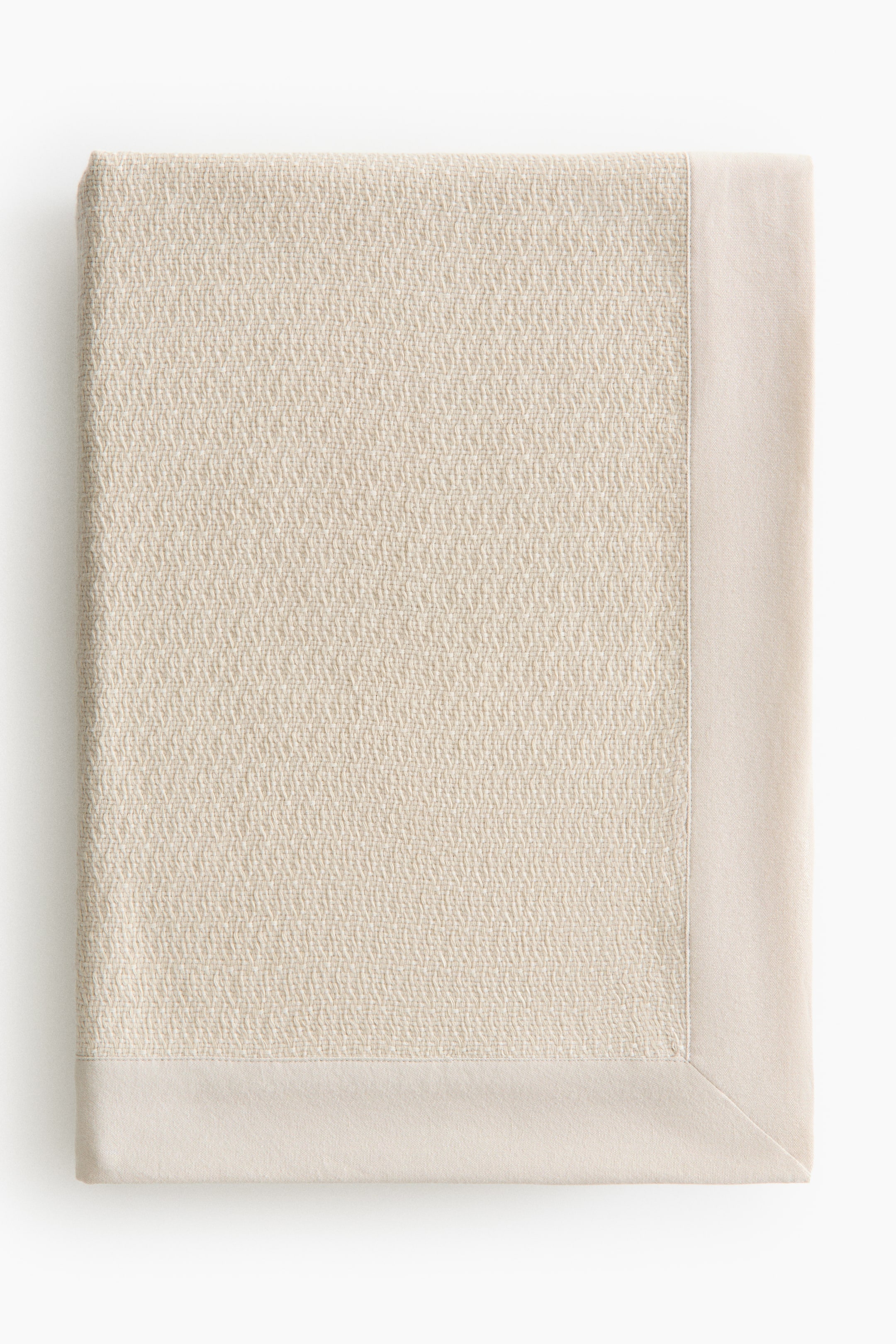 View larger image: A light beige blanket is neatly folded into a rectangle, showcasing a textured weave across its main body. It is bordered by a smooth, plain edge visible along the right and bottom sides.