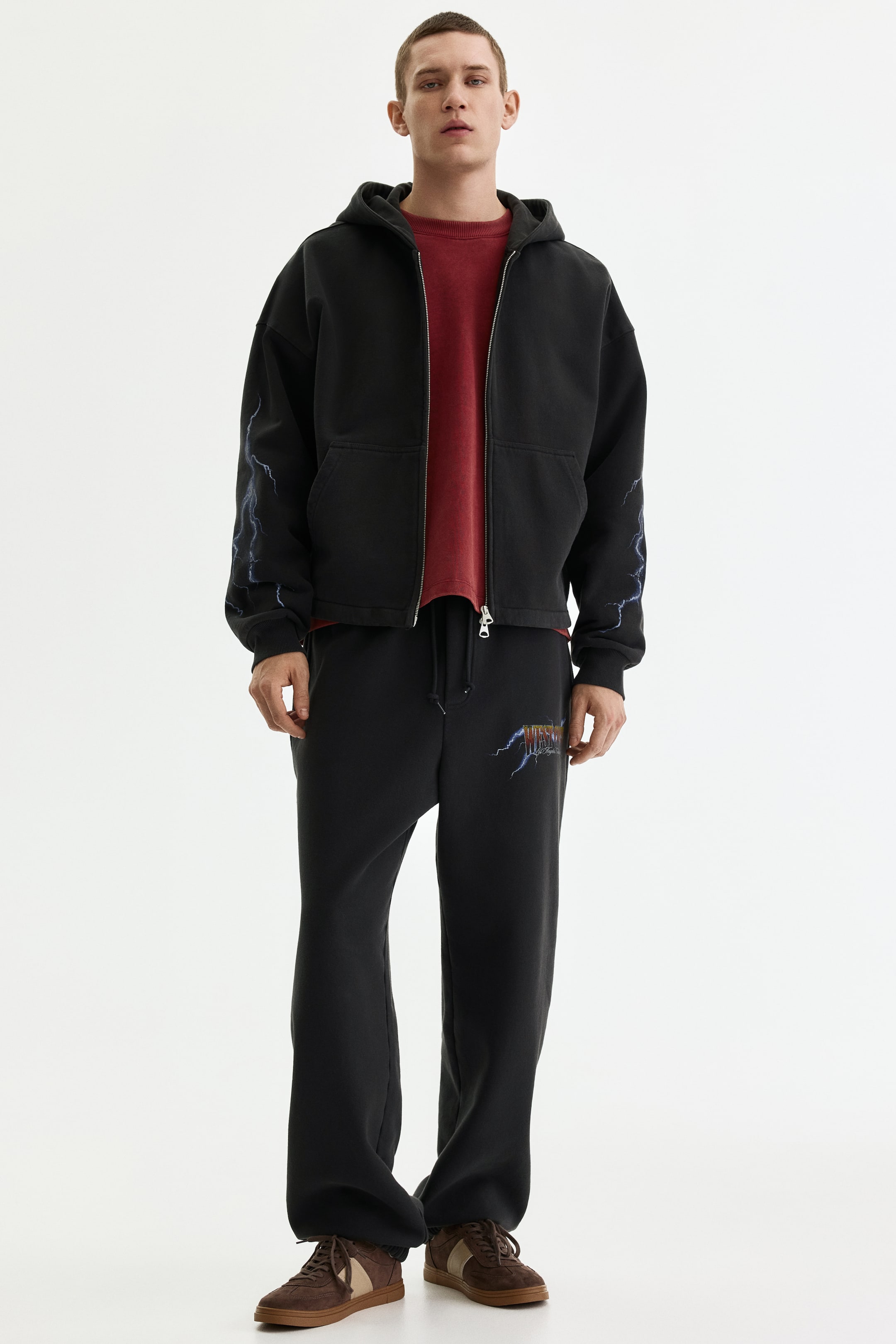 View larger image: A black oversized zip-up hoodie features a drawstring hood, a full-zip closure, two front patch pockets, and blue lightning bolt graphics on both sleeves. It is paired with matching black sweatpants that feature an elastic waistband with a drawstring, a relaxed straight-leg fit, and a colourful lightning bolt graphic on the upper left thigh.