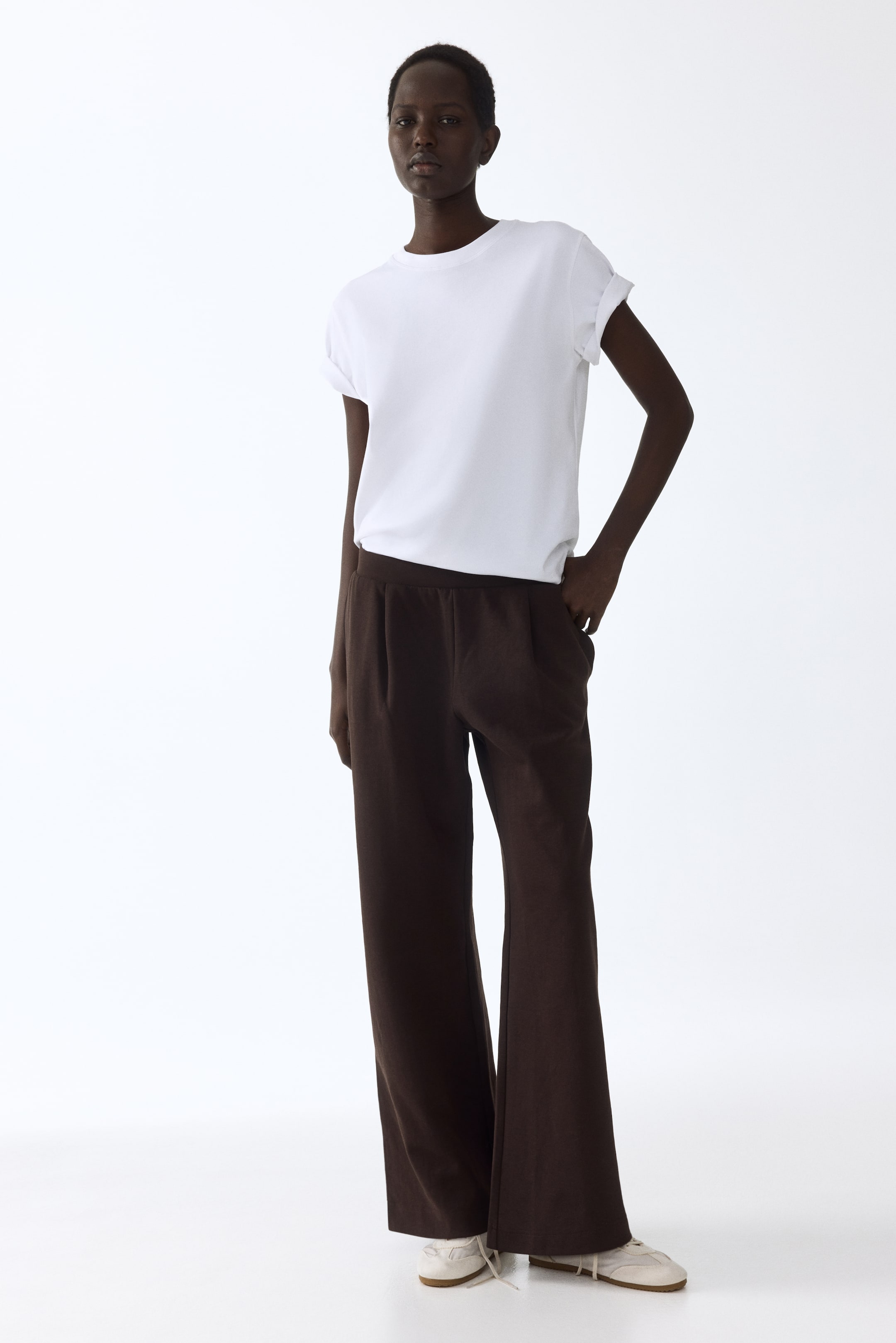 הצגת תמונה גדולה יותר: חולצת טי לבנה עם שרוולים קצרים ושרוולים מגולגלים עם צווארון עגול נלבשת, תחובה בתוך מכנסיים רחבים בצבע חום כהה. המכנסיים כוללים גומי במותן, כיסים בצדדים וקפלים בחזית.