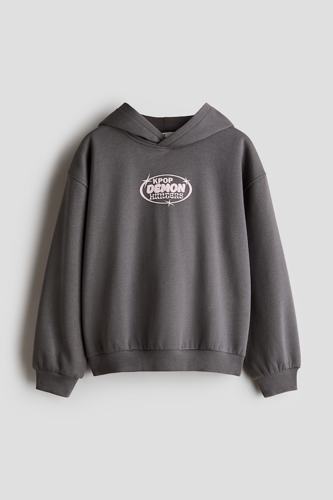 A dark gray pullover hoodie is hanging in a front-facing view, displaying a light pink oval graphic on the center chest that reads "KPOP DEMON HUNTERS" with star accents. The hoodie is featuring drop shoulders, long sleeves with ribbed cuffs, and a ribbed hem. - 1