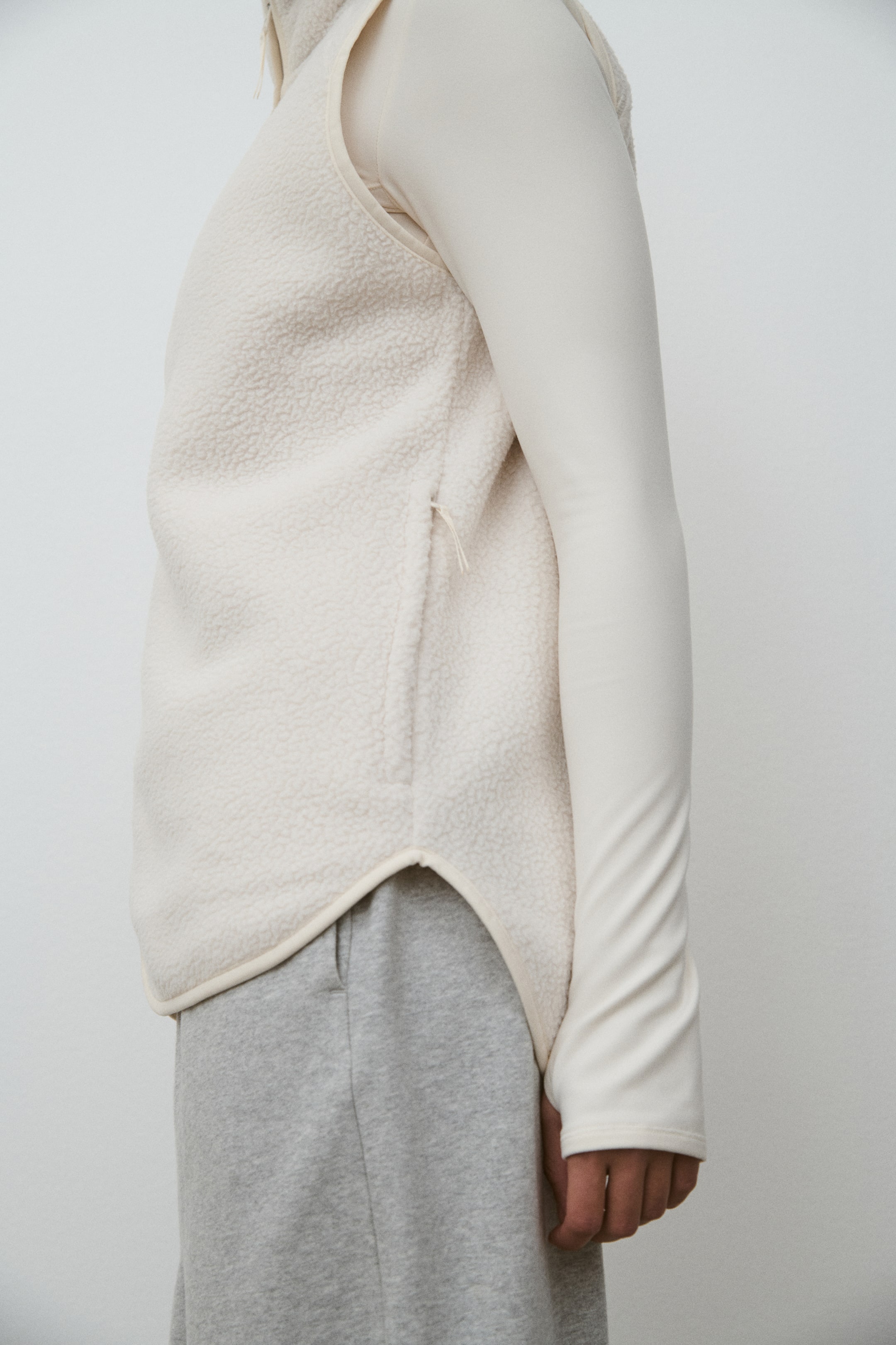 View larger image: A cream teddy sports gilet is worn by the model, layered over a long-sleeved top in a similar color. It features a zippered side pocket, elastic binding along the armholes and hem, and a rounded hemline.
