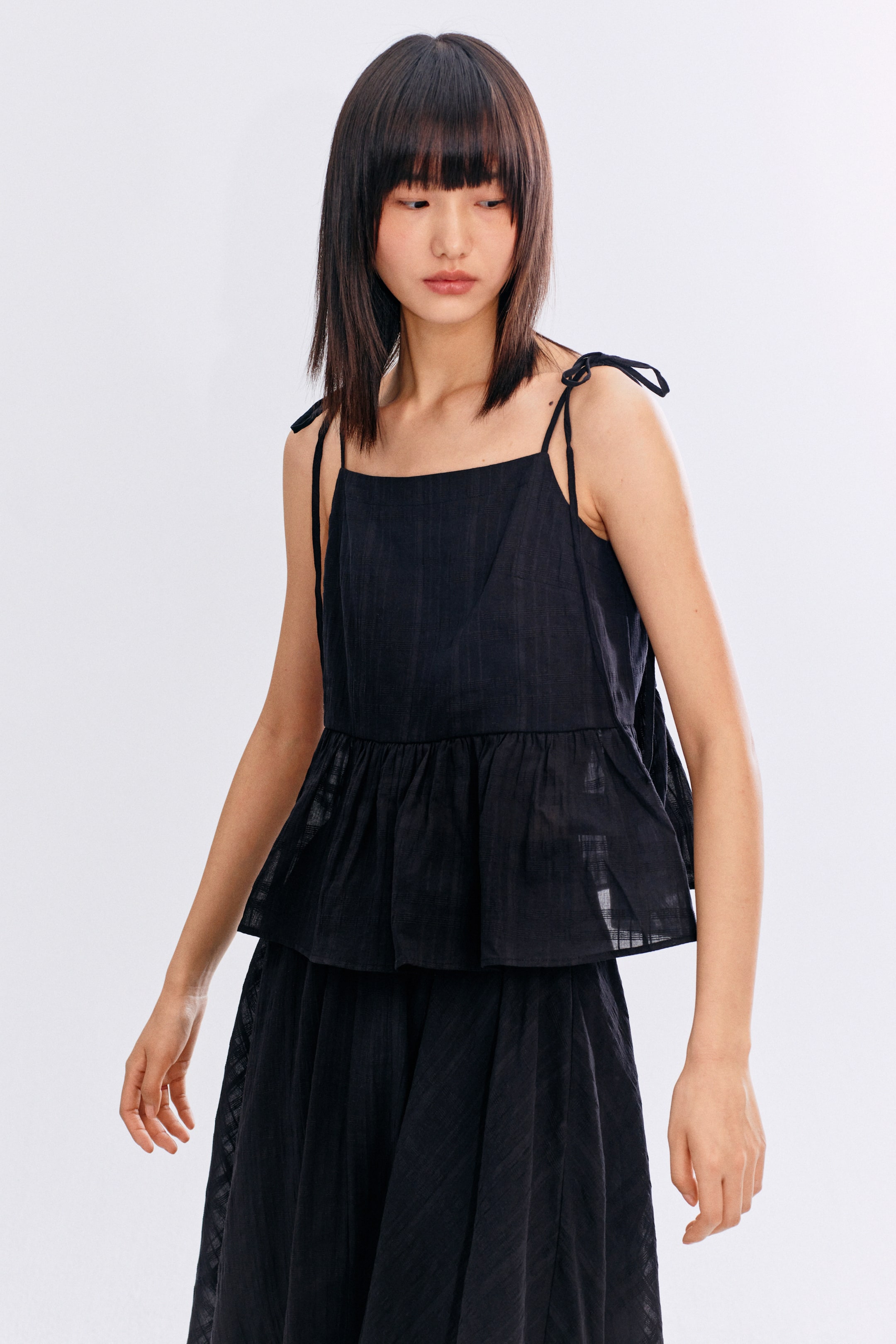 拡大画像を表示: 黒のキャミソールトップは、スクエアネックライン、肩紐をリボン結びにしたデザイン、ギャザー入りのペプラムヘムが特徴で、同じく黒のスカートと合わせています。どちらも微妙なテクスチャーのストライプ模様が施されています。