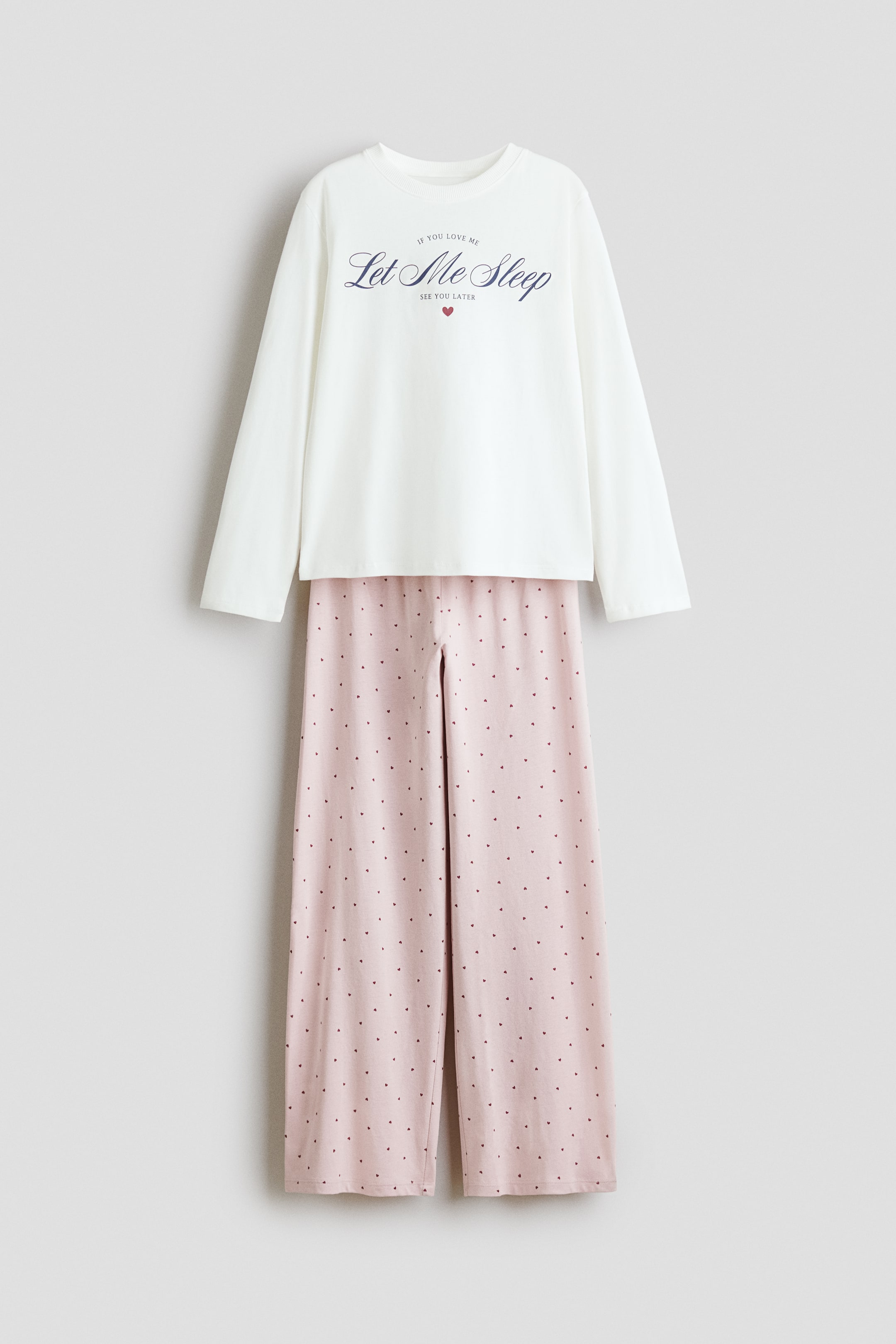 Größeres Bild anzeigen: Ein Pyjama-Set besteht aus einem weißen Langarmshirt mit Rundhalsausschnitt und einer pinkfarbenen Hose mit weitem Bein. Das Oberteil zeigt den Text "IF YOU LOVE ME Let Me Sleep SEE YOU LATER" mit einem kleinen roten Herz, während die Hose ein Allover-Muster aus kleinen dunklen herzförmigen Punkten aufweist.
