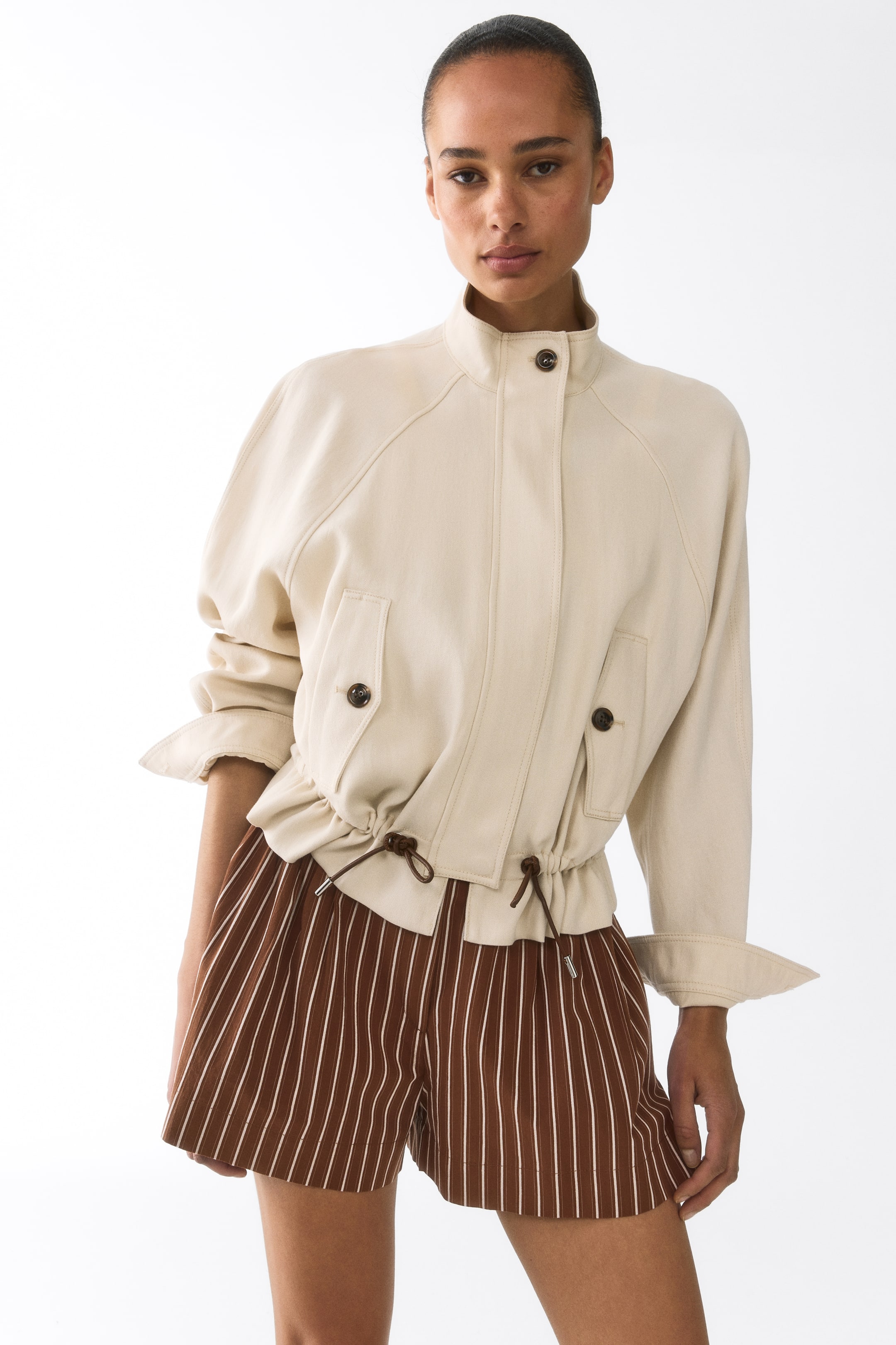 Agrandir l'image: Une veste crème, vue sous un angle légèrement de face à droite, présente un col montant avec une fermeture à bouton visible en haut et des manches raglan retroussées aux poignets. Elle est conçue avec deux poches à rabat à l'avant avec boutons, et un cordon de serrage resserre la taille, créant un ourlet froncé.