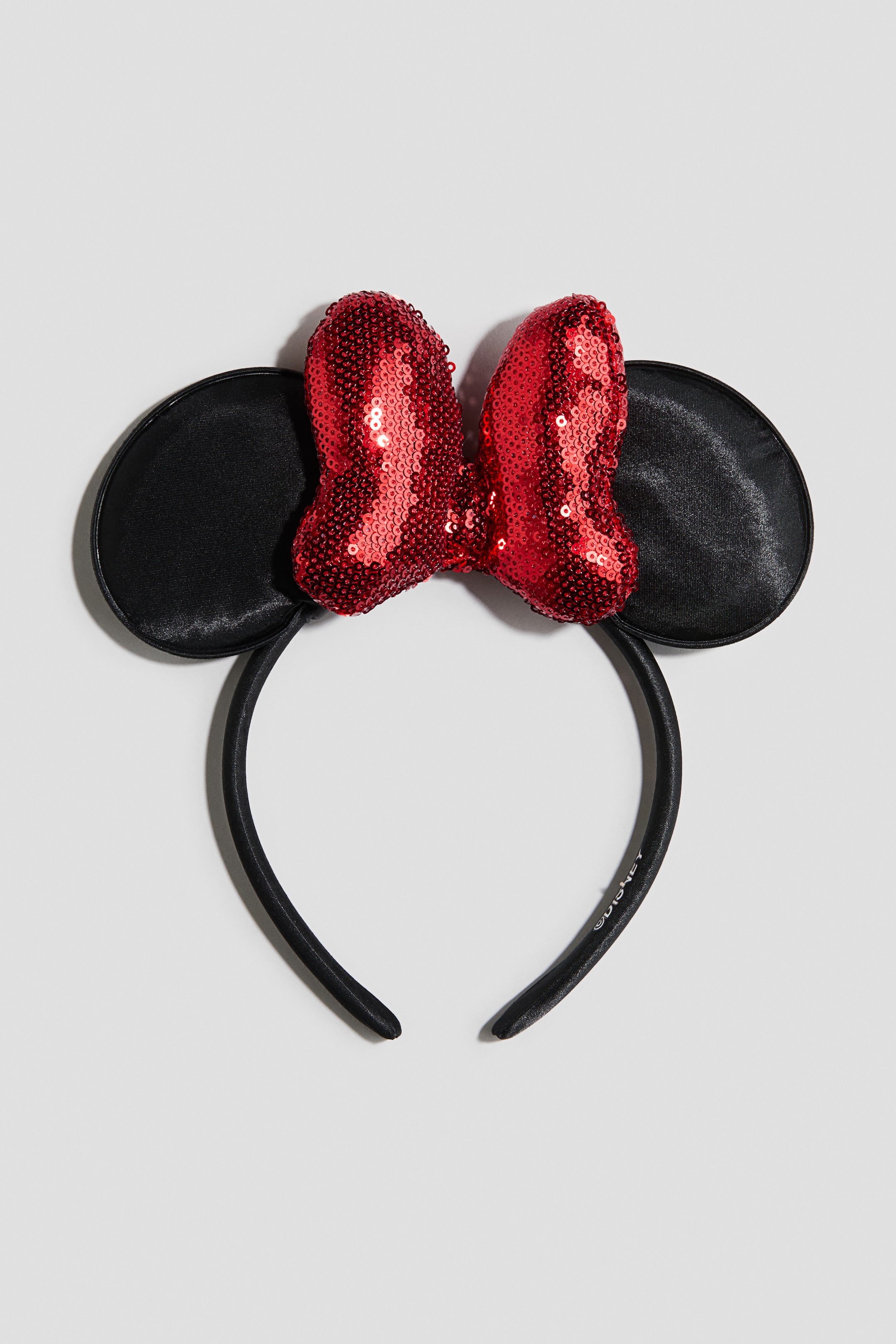 Zobrazit větší obrázek: Čelenka s mašlí je zobrazena zepředu a je tvořena černou páskou potaženou látkou, ke které jsou připevněny dvě kulaté černé saténové ouška. Uprostřed mezi oušky spočívá velká polstrovaná červená mašle pokrytá třpytivými flitry.