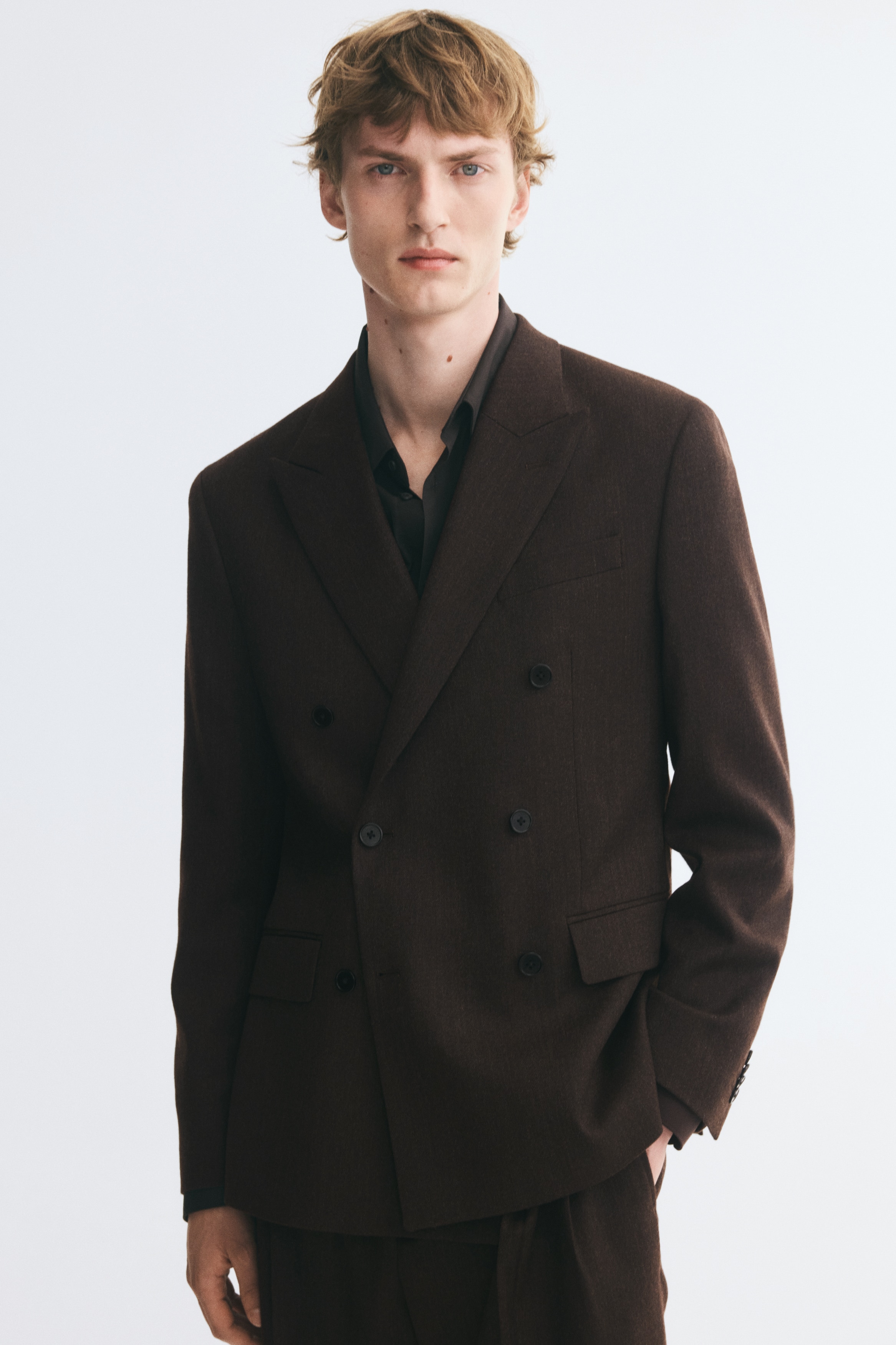 A dark brown, double-breasted blazer is shown from the front, featuring wide peak lapels, a six-button front fastening, and two lower flap pockets. The long sleeves end in buttoned cuffs, and a single welt pocket appears on the left chest.