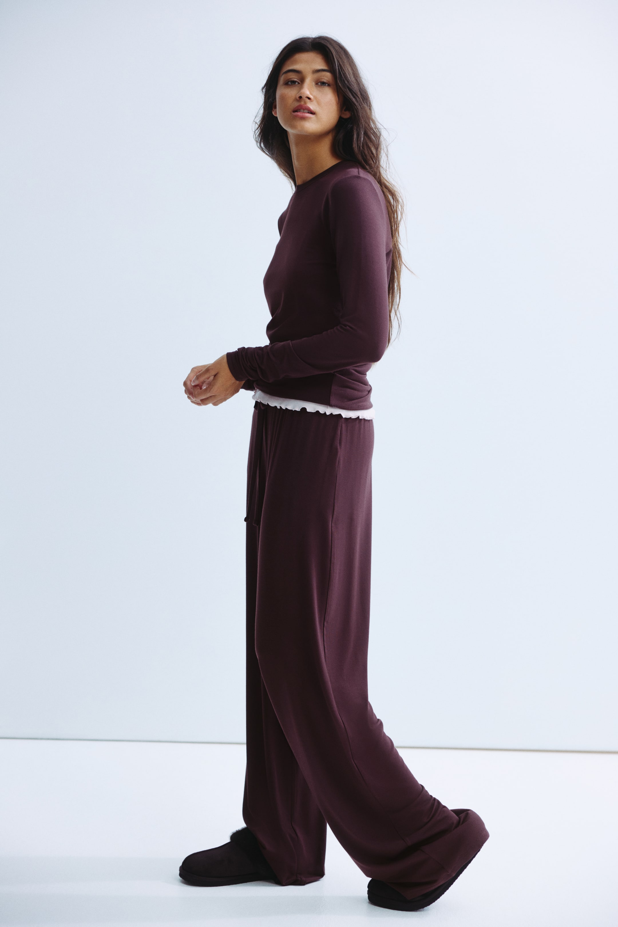 Grotere afbeelding bekijken: Een donker kastanjebruine top met lange mouwen en bijpassende wijduitlopende broek worden van opzij gepresenteerd. De top heeft een ronde hals en sluit nauw aan, met een witte gerimpelde boord die zichtbaar aan de taille tevoorschijn komt. De broek valt losjes en heeft een tailleband met trekkoord.