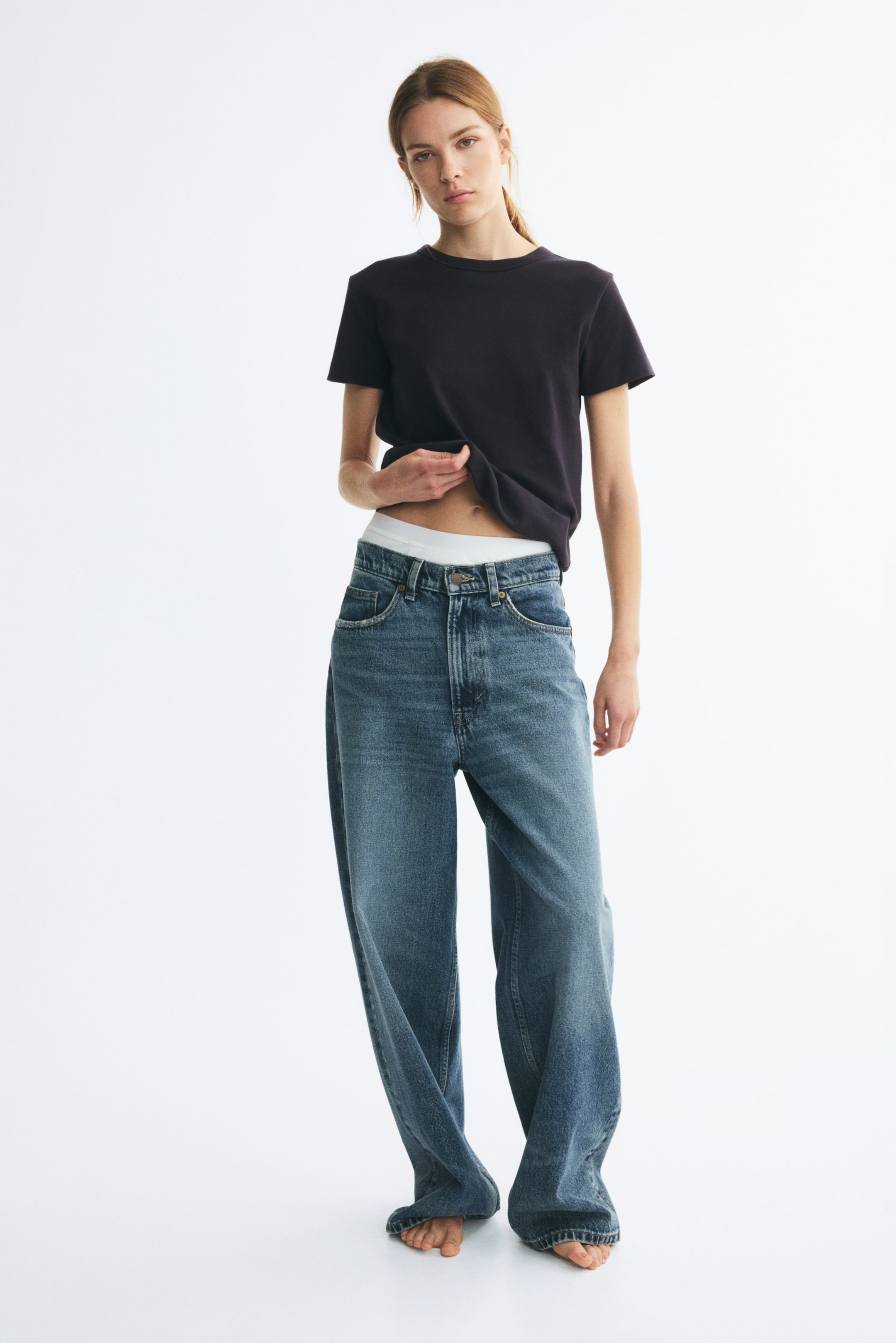 拡大画像を表示: モデルは、厚手のコットンジャージー素材のダークカラーのフィットTシャツを着用しており、前裾を少し持ち上げて下に白いウエストバンドを見せています。半袖Tシャツはリブ仕様のクルーネックで、ワイドレッグのブルージーンズと合わせています。