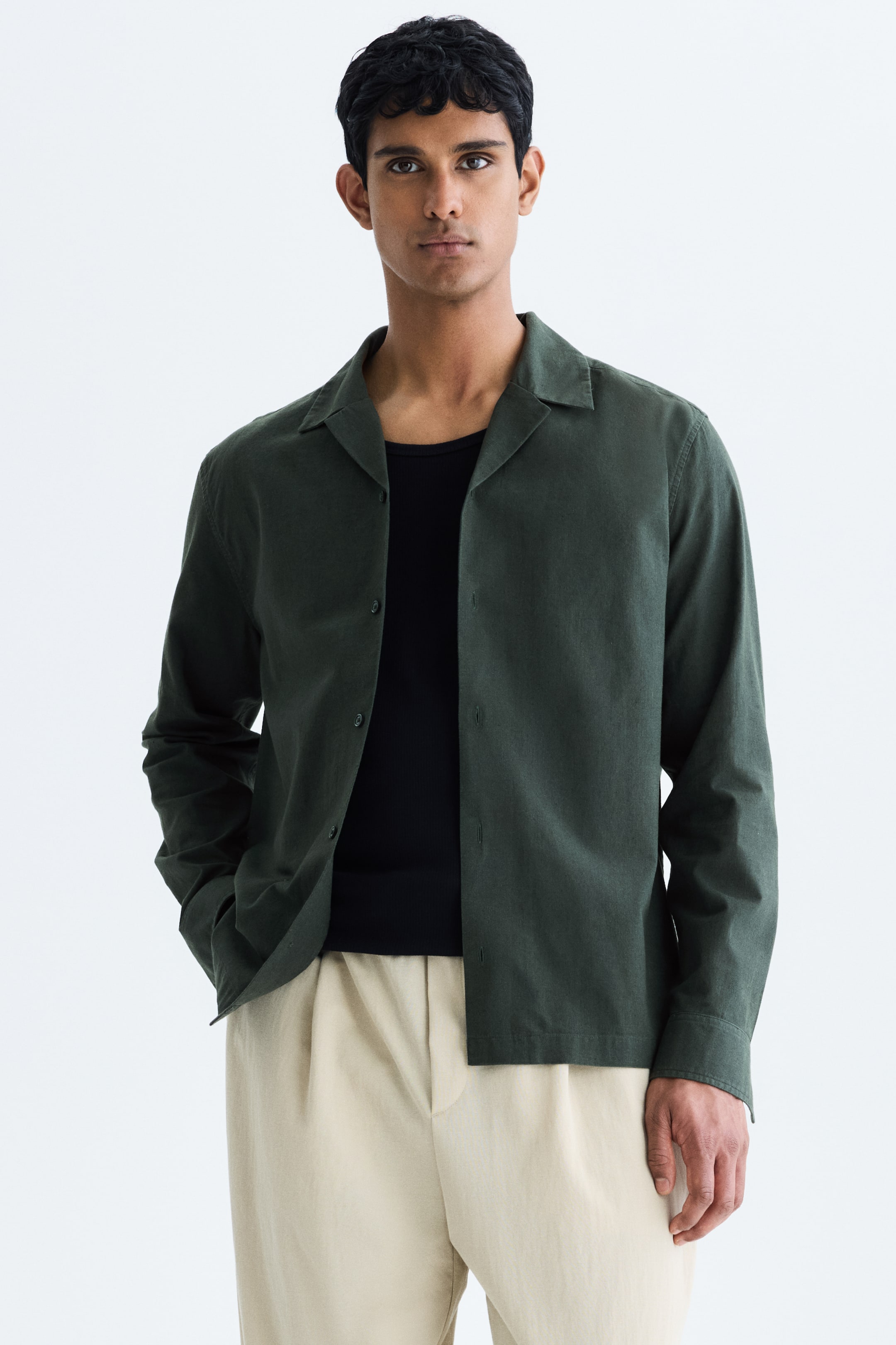 View larger image: The model is wearing a dark green long-sleeved shirt with a textured weave, resort collar, and visible front buttons, open over a black undershirt. This relaxed-fit shirt comes with long sleeves, unbuttoned cuffs, and a straight hem, paired with beige pleated pants; the model's hand rests casually in a pant pocket.