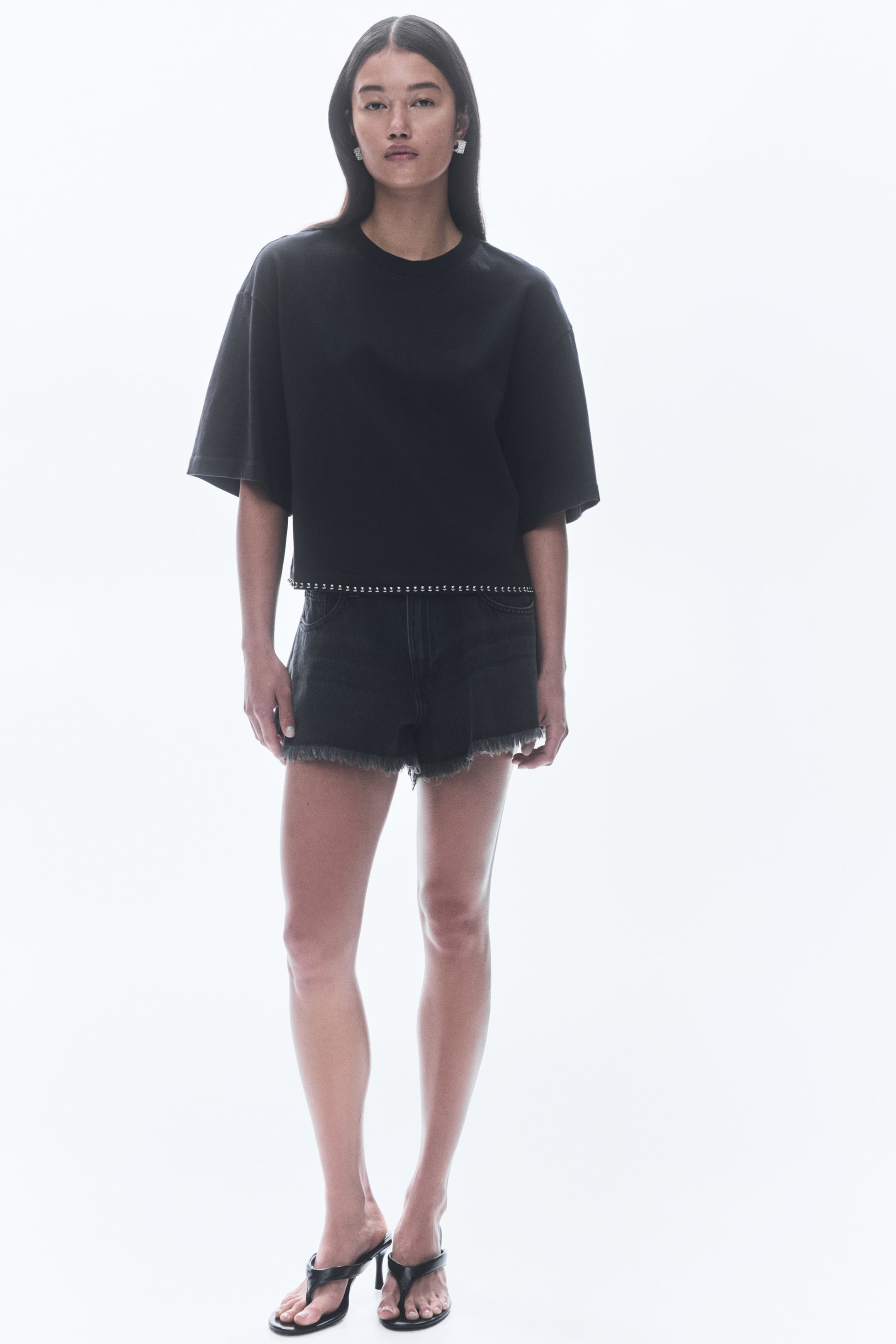 View larger image: A black cropped t-shirt, facing forward, is featuring a crew neck and wide short sleeves. Its straight hem is detailed with a line of silver studs, and it is being styled with dark wash denim shorts showcasing a frayed hem.