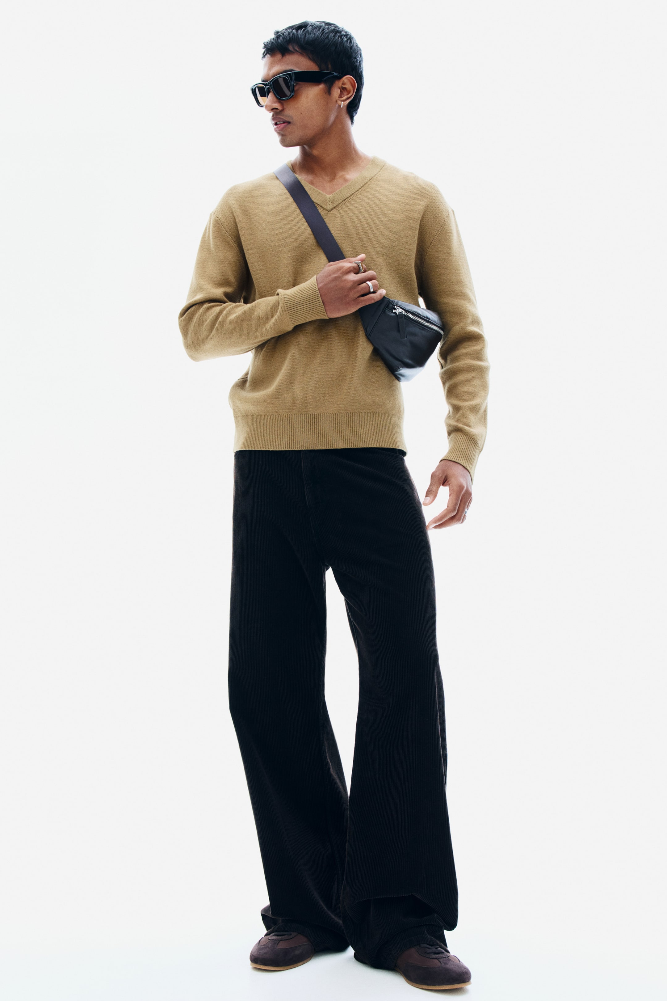 View larger image: A beige cotton-blend knit sweater featuring a ribbed V-neck, long sleeves, and ribbed trim at the cuffs and hem is worn by the model. The relaxed-fit sweater is styled with black wide-leg trousers and a black leather crossbody bag worn across the chest.