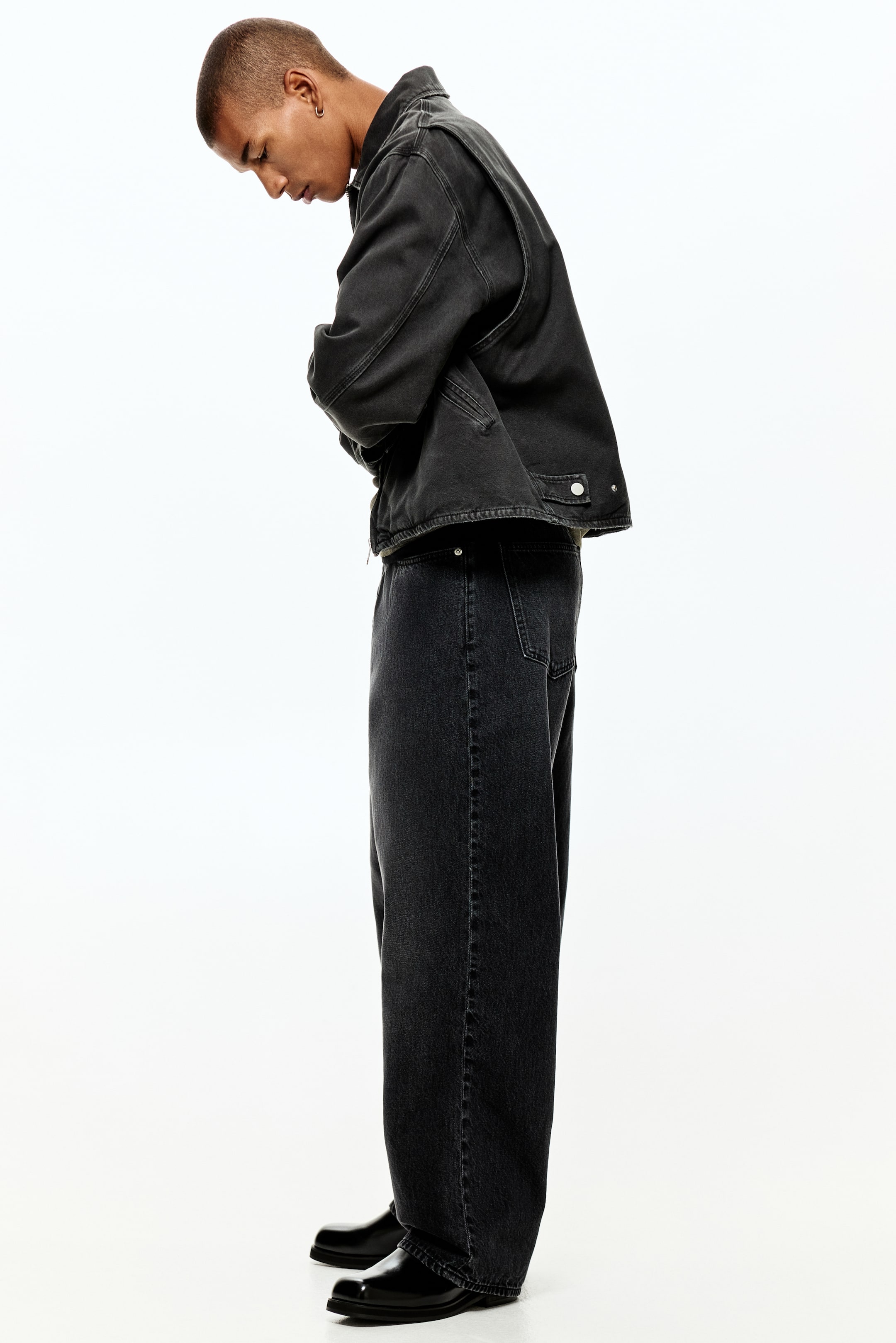 Ampliar la imagen: Una chaqueta de mezclilla negra lavada y jeans de pierna ancha se presentan desde un perfil lateral. La chaqueta presenta un cuello en punta, mangas largas y cierres de botones plateados en los puños y la pretina. Los jeans sueltos de pierna ancha caen sobre zapatos negros pulidos.