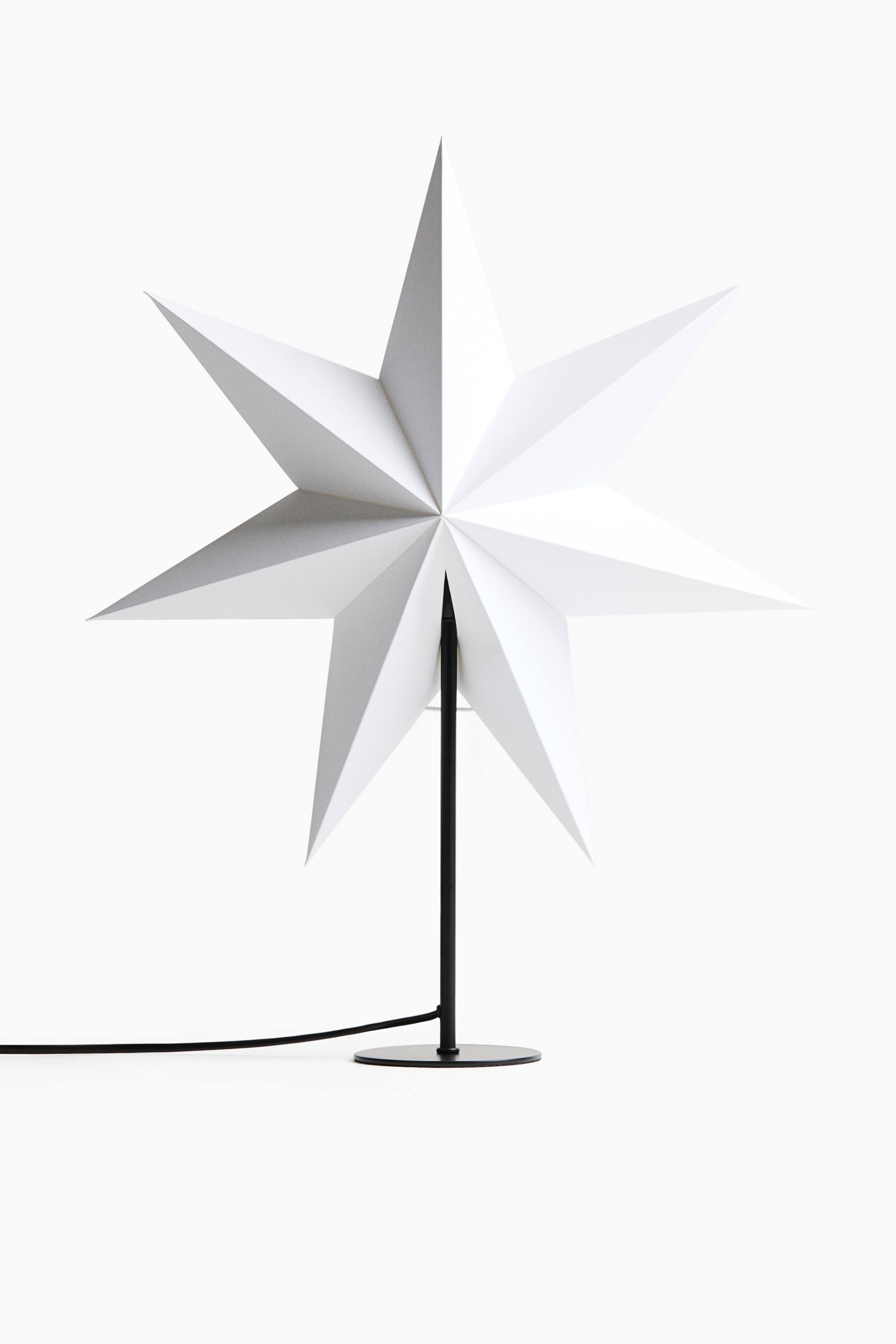View larger image: The table lamp features a large, white, seven-pointed folded paper star shade, shown front-facing on a slender black metal stand. The stand rises from a flat, circular black base, with a black power cord extending to the left.