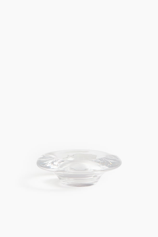 Un portavelas de vidrio transparente presenta una parte superior amplia y facetada que se ensancha hacia afuera y curva hacia abajo hasta una base sólida y escalonada. Su superficie lisa y transparente capta y refleja la luz ambiental. - 1