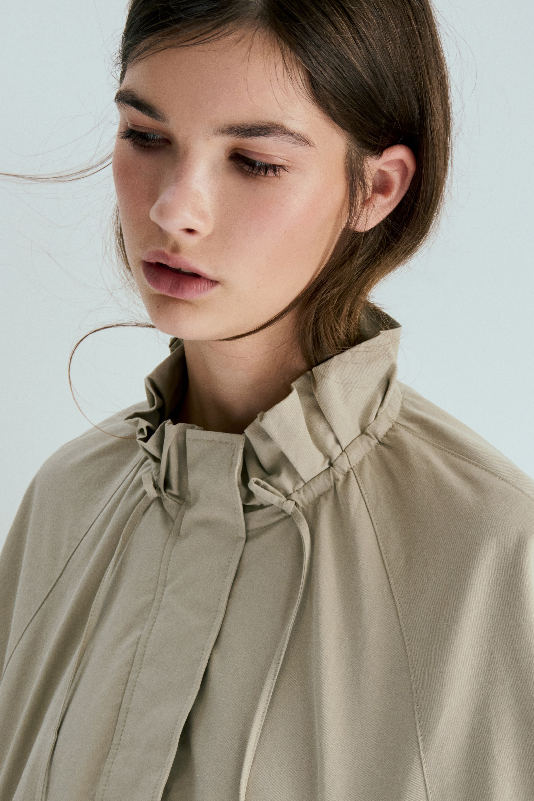 View larger image: A beige jacket features a high, gathered stand-up collar with a noticeable drawstring and ties at the front. Raglan sleeves extend from the collar seam, and a placket covers the front closure.