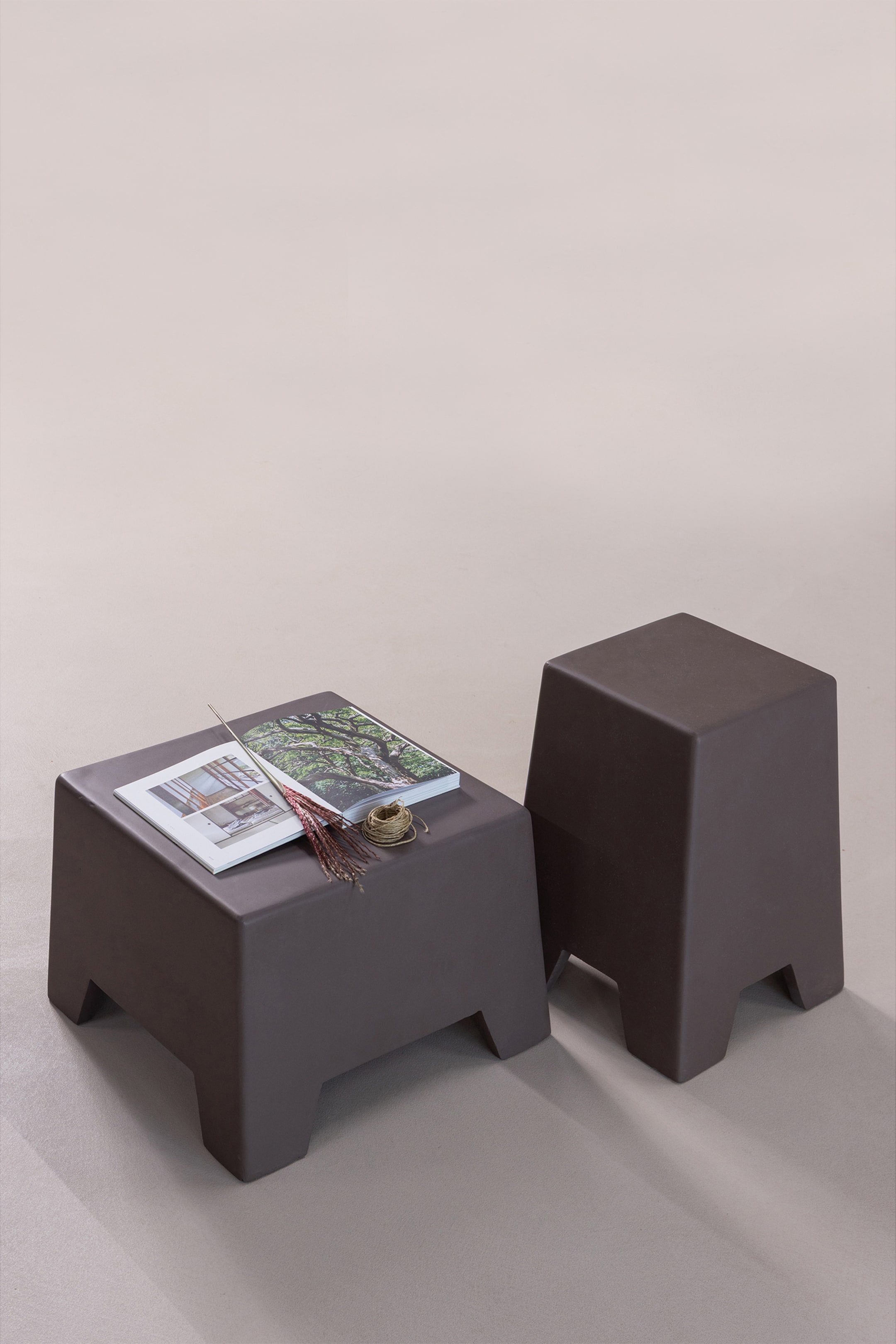 Größeres Bild anzeigen: Zwei dunkelbraune Hocker mit quadratischen Oberflächen, sich verjüngenden Seiten und integrierten Blockbeinen. Der kürzere Hocker, der im Vordergrund schräg steht, präsentiert eine Zeitschrift, eine getrocknete Pflanze und eine Schnur auf seiner Oberseite, während der höhere Hocker dahinter steht.