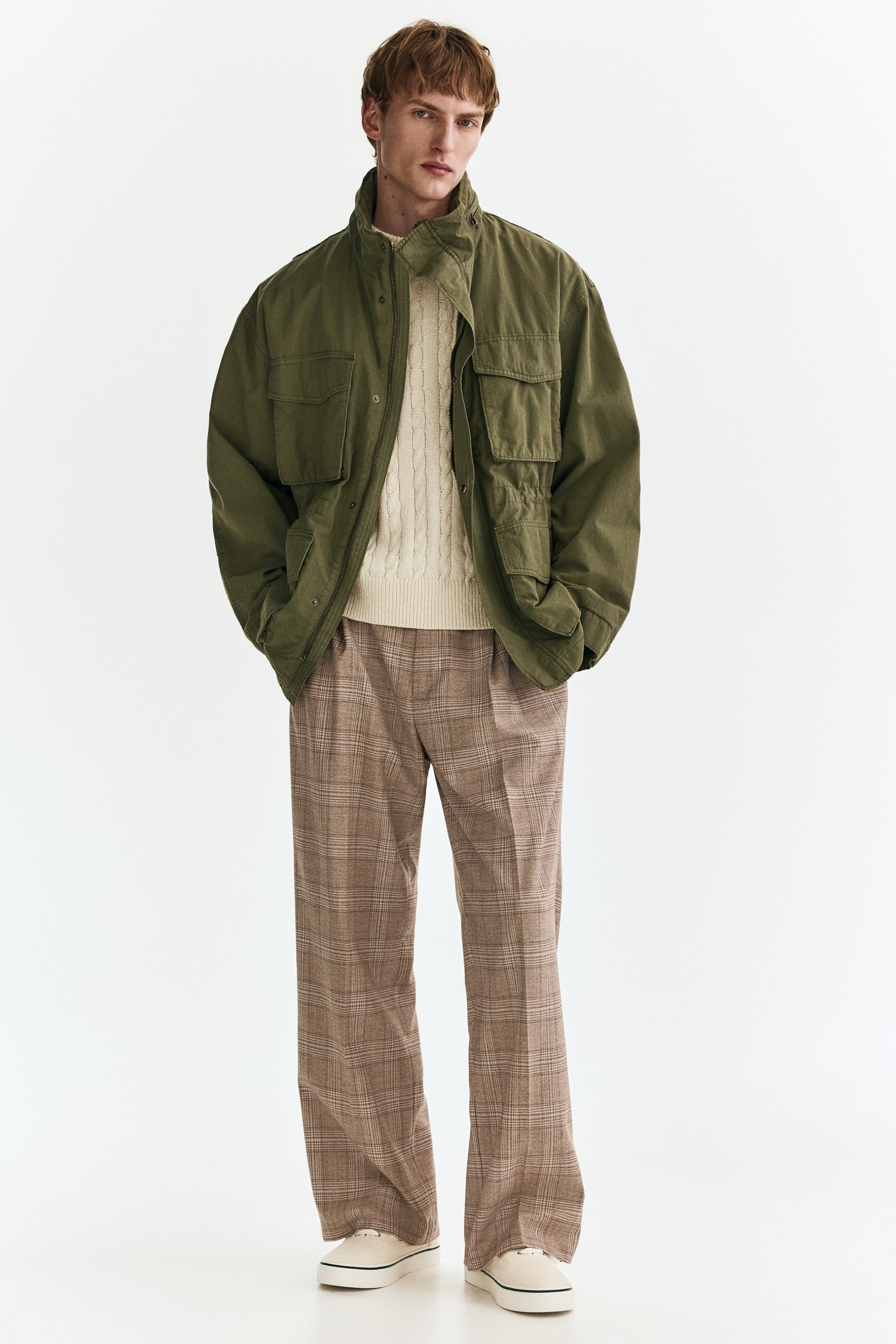 Visa större bild: En olivgrön utilityjacka visas framifrån, med en ståkrage, en fullständig dragkedjestängning med en tryckknappslå och fyra fickor med lock på framsidan. Den bärs öppen över en krämfärgad kabelstickad tröja, vilket framhäver dess avslappnade passform och markerade midja.