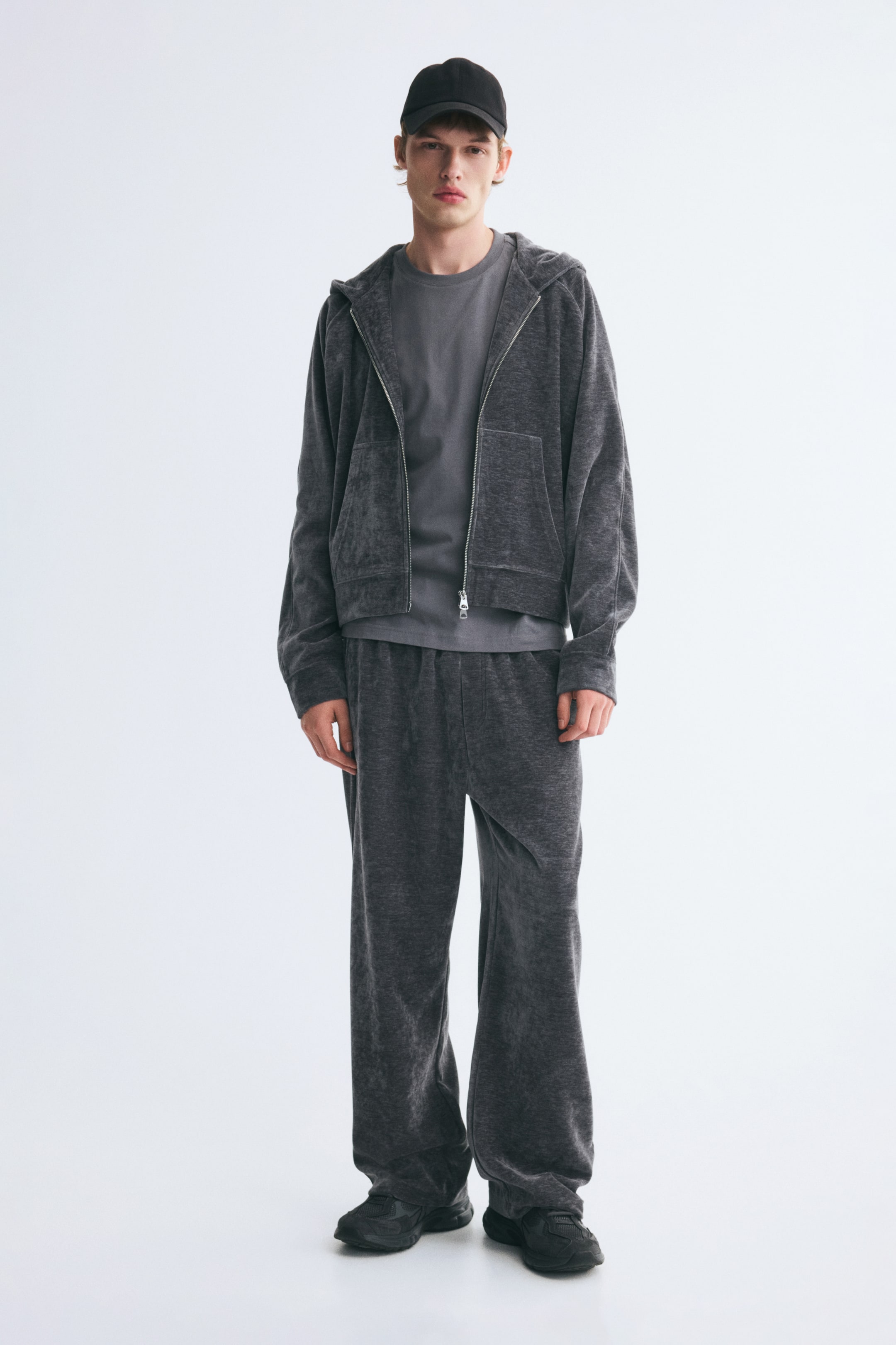 View larger image: A dark grey velour tracksuit, featuring a zip-up hooded jacket and matching wide-leg trousers, is worn by the model. The jacket is styled open over a light grey crewneck T-shirt, and the outfit is completed with a black baseball cap and black sneakers.
