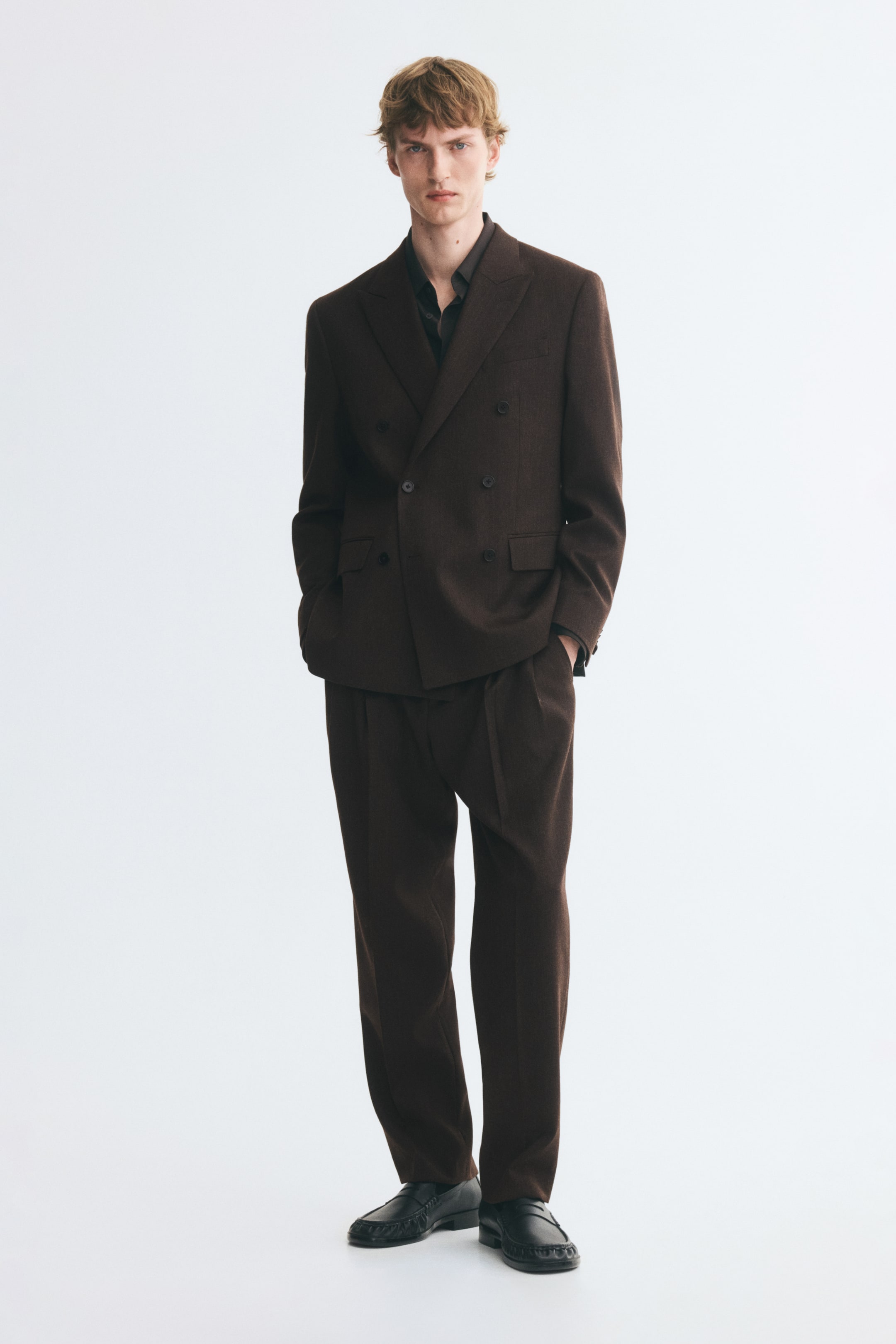 View larger image: Dark brown wool suit pants featuring a regular fit, front pleats, and pressed creases are worn by the model. The model is standing with one hand resting in a diagonal side pocket, styling the pants with a matching double-breasted blazer and black leather loafers.