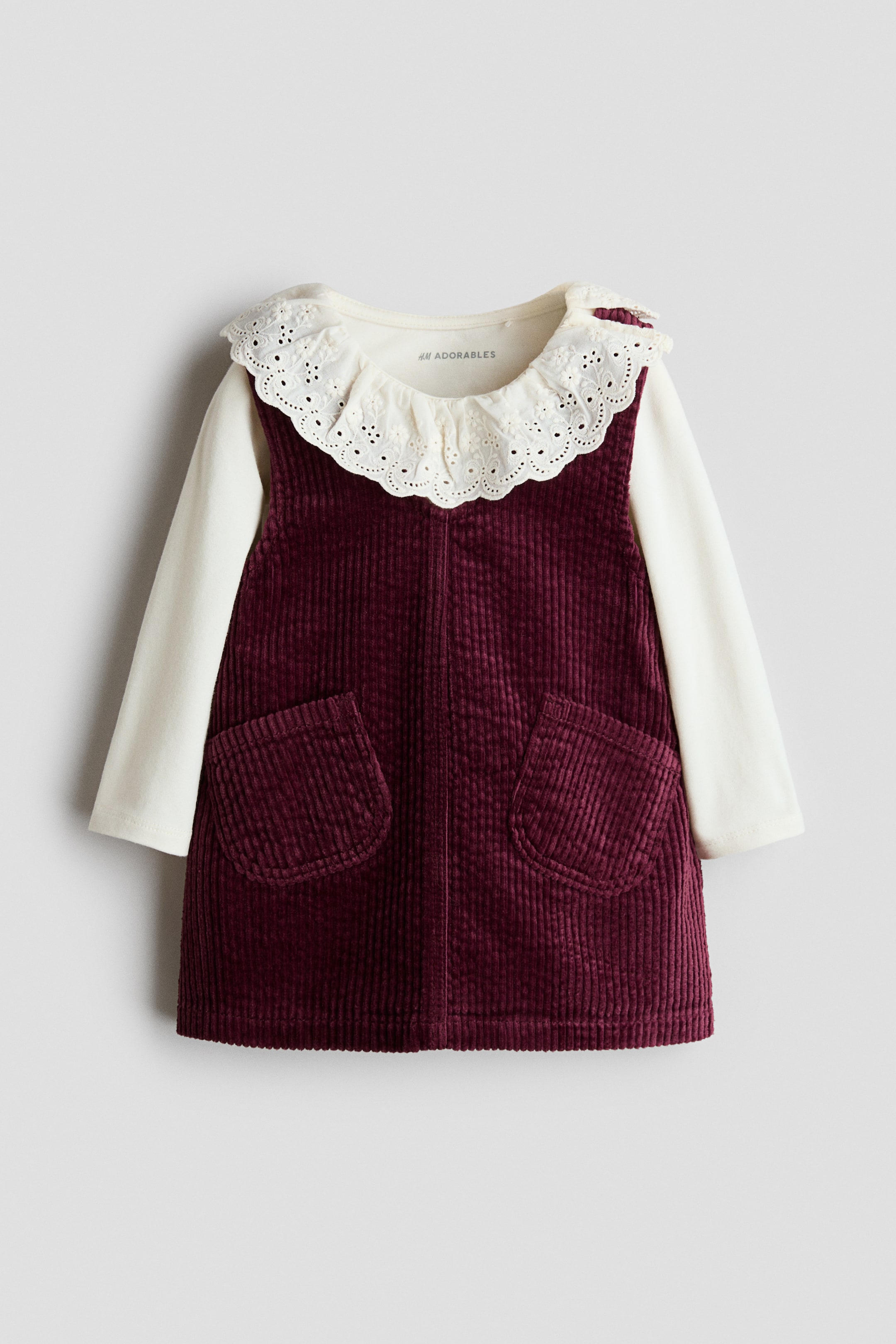 Προβολή μεγαλύτερης εικόνας: Ένα σετ H&M Adorables παρουσιάζει πρόσοψη ενός μπορντό κοτλέ φορέματος pinafore, με λαιμόκοψη σε σχήμα V και δύο μπροστινές εξωτερικές τσέπες. Φοριέται πάνω από ένα κρεμ τοπ με μακριά μανίκια, που διαθέτει γιακά με scalloped broderie anglaise και κουμπώματα πίεσης στον έναν ώμο.