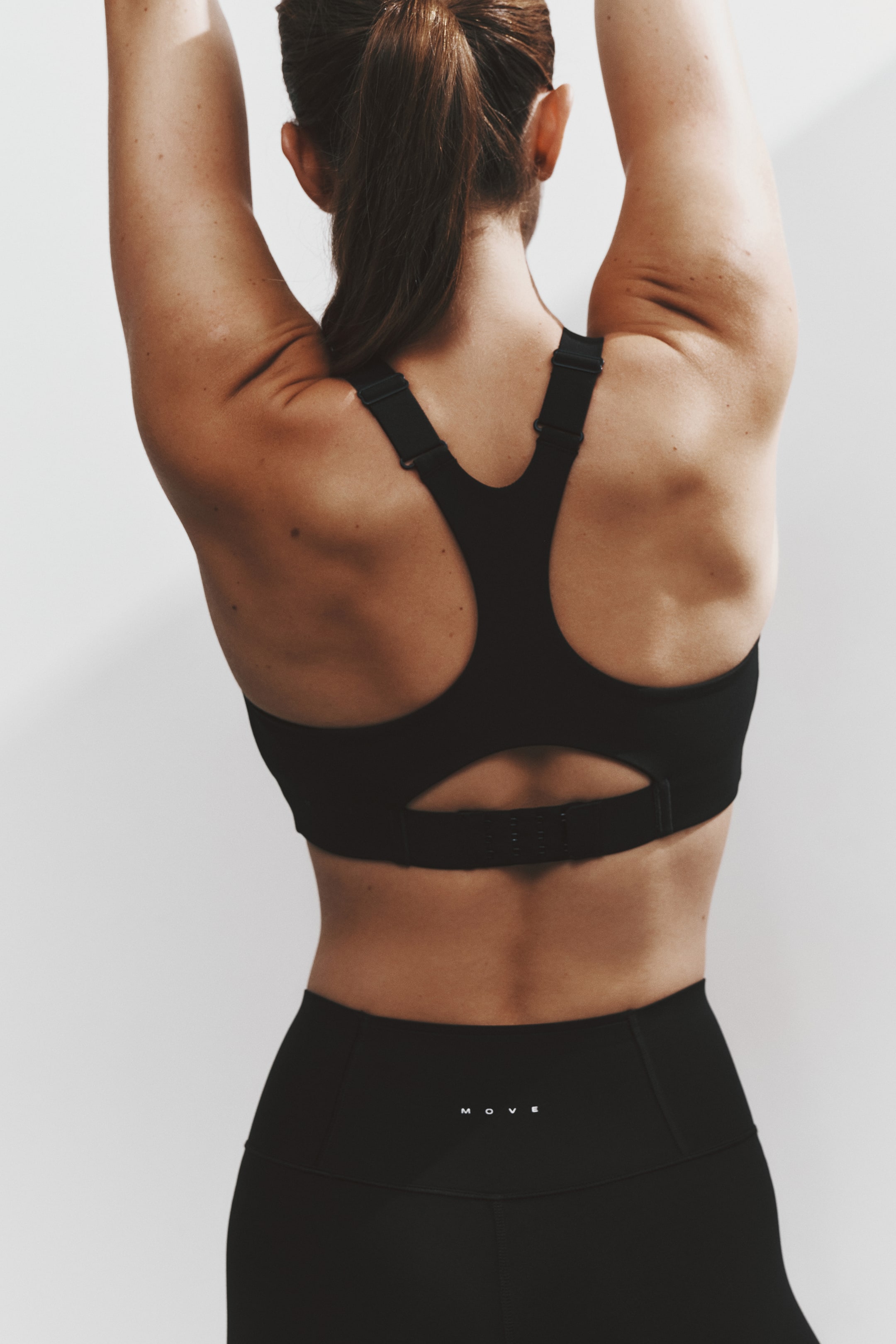 View larger image: A black sports bra is shown from the back, featuring a racerback silhouette with adjustable shoulder straps, a lower back cutout, and a hook-and-eye band closure. It is paired with high-waisted black leggings featuring a small white "MOVE" logo centered on the back waistband.