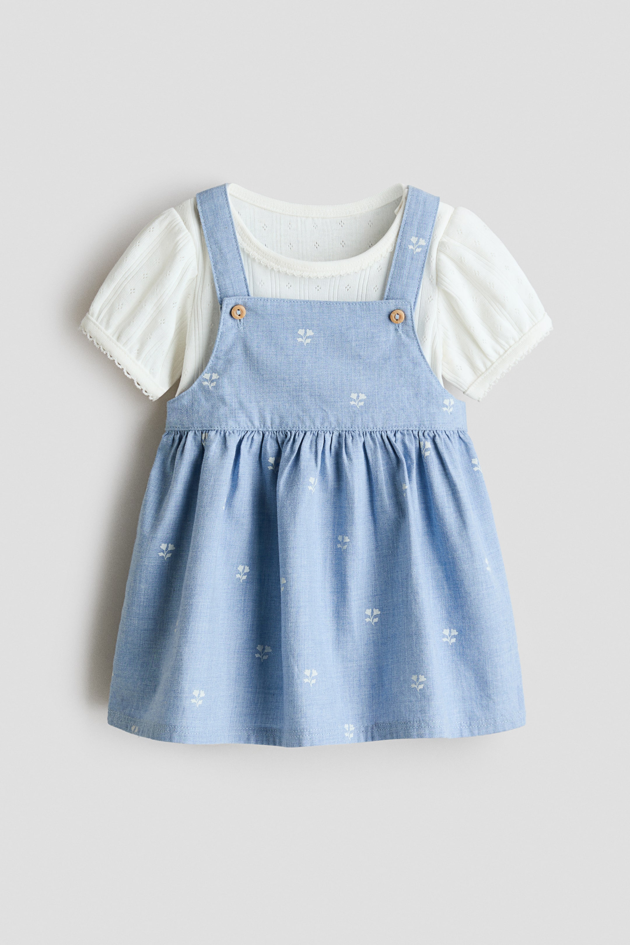 Agrandir l'image: Une robe salopette bleu clair, ornée de motifs floraux blancs et d'une taille froncée, est superposée sur un haut blanc à manches courtes, présentée de face. La robe salopette est dotée de boutons en bois sur ses bretelles de bavette avant, et le haut blanc arbore un col rond bordé de dentelle festonnée et des manches bouffantes.