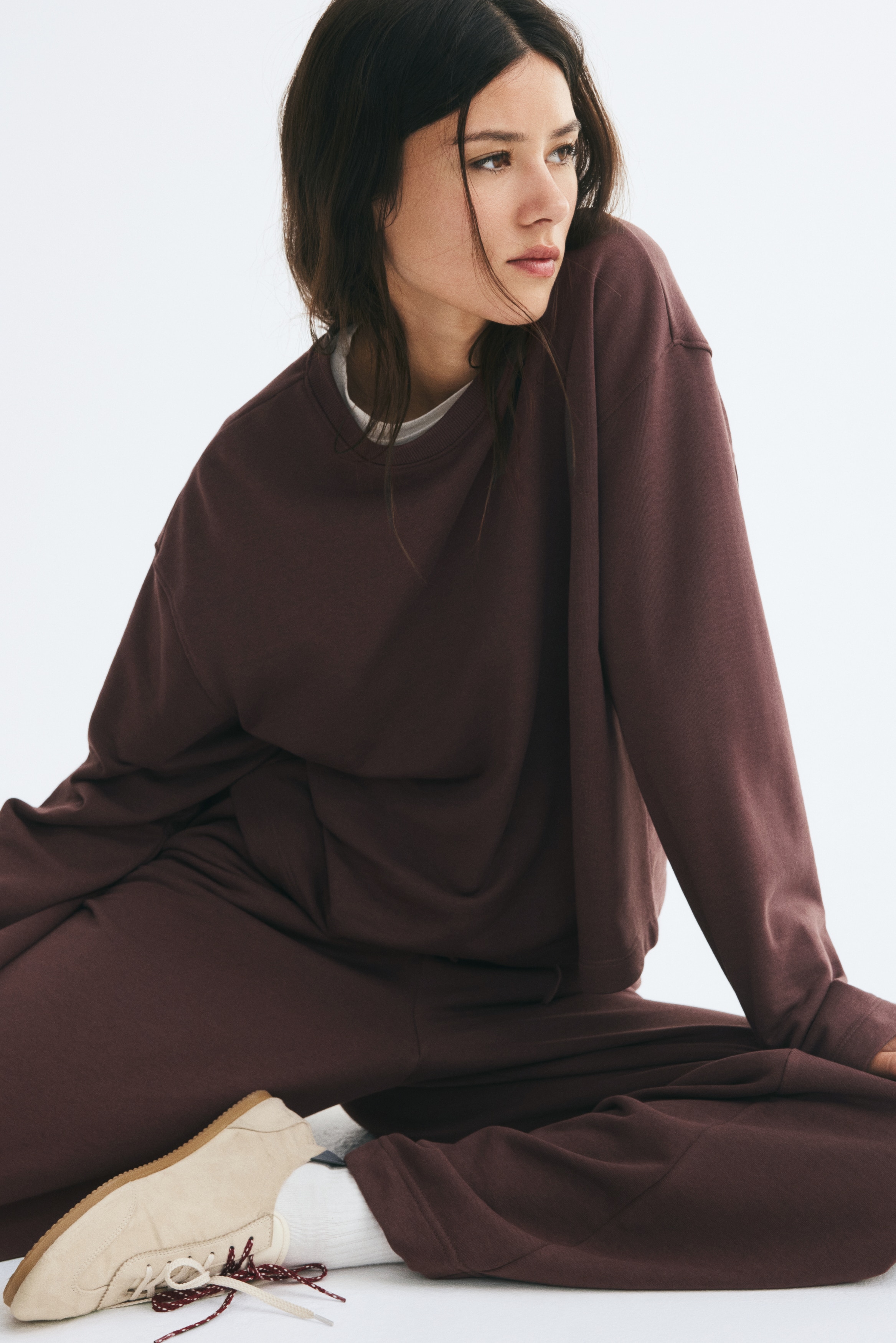 Een donkerbruin tweedelig setje wordt frontaal gepresenteerd, bestaande uit een top met lange mouwen en ronde hals en bijpassende rechte broek. De top met een relaxte pasvorm valt over de broek, die een elastische tailleband heeft.