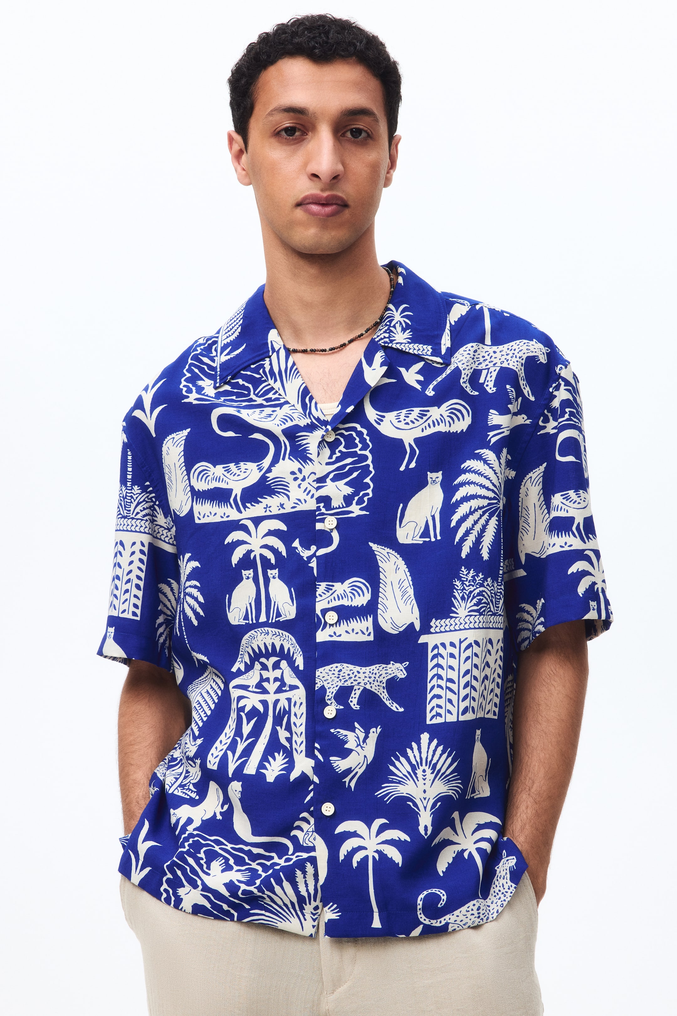 View larger image: The model dons a short-sleeved shirt in a regal blue printed weave, showcasing a white tropical motif of flora and fauna. This shirt boasts a resort collar, a French front, a relaxed fit, and a straight-cut hem. The top buttons are left open, revealing a beaded necklace.