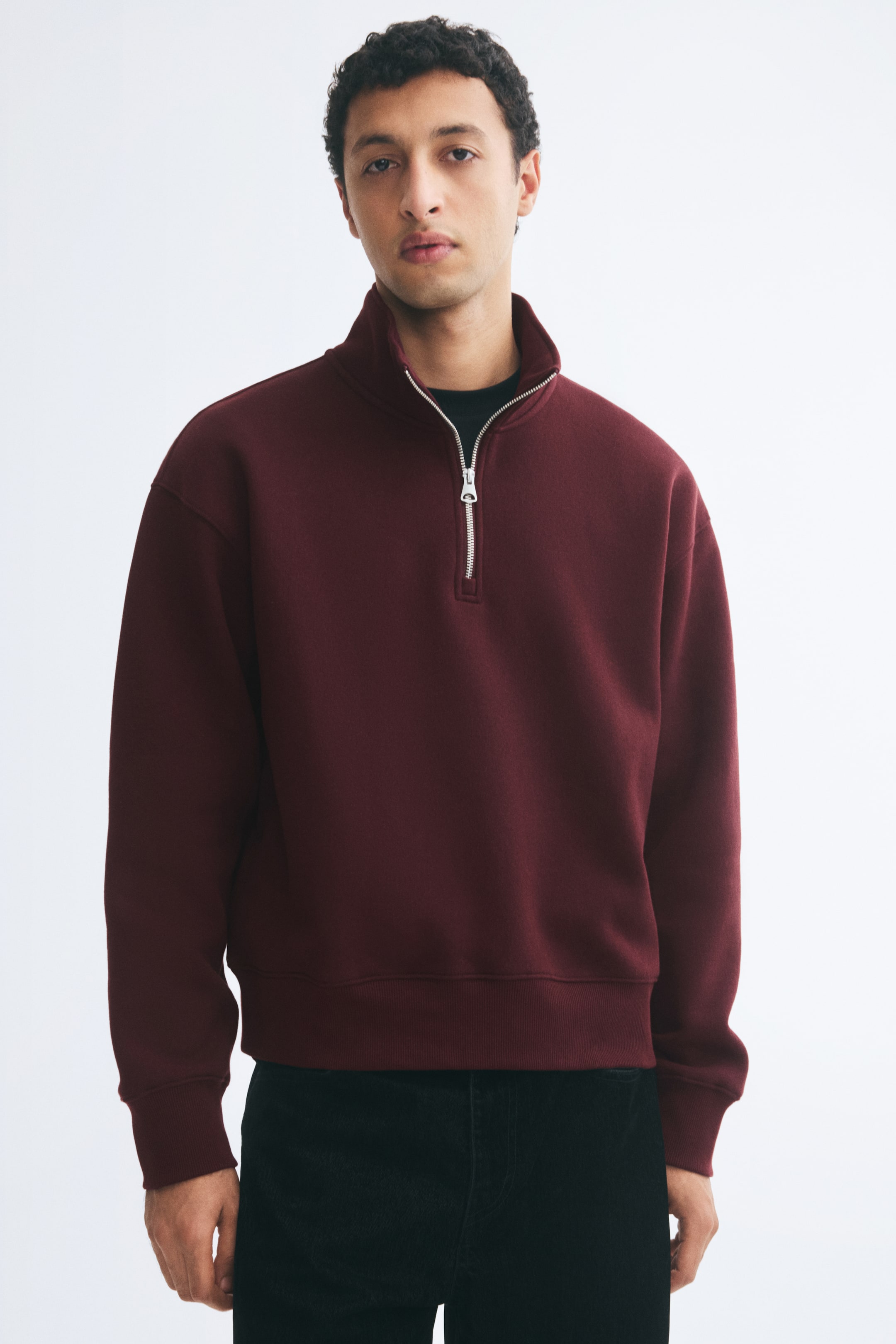 View larger image: The model is wearing a burgundy heavyweight half-zip sweatshirt featuring a stand collar, dropped shoulders, and ribbed cuffs and hem. The relaxed-fit jumper is layered over a dark crew-neck T-shirt and paired with black trousers.