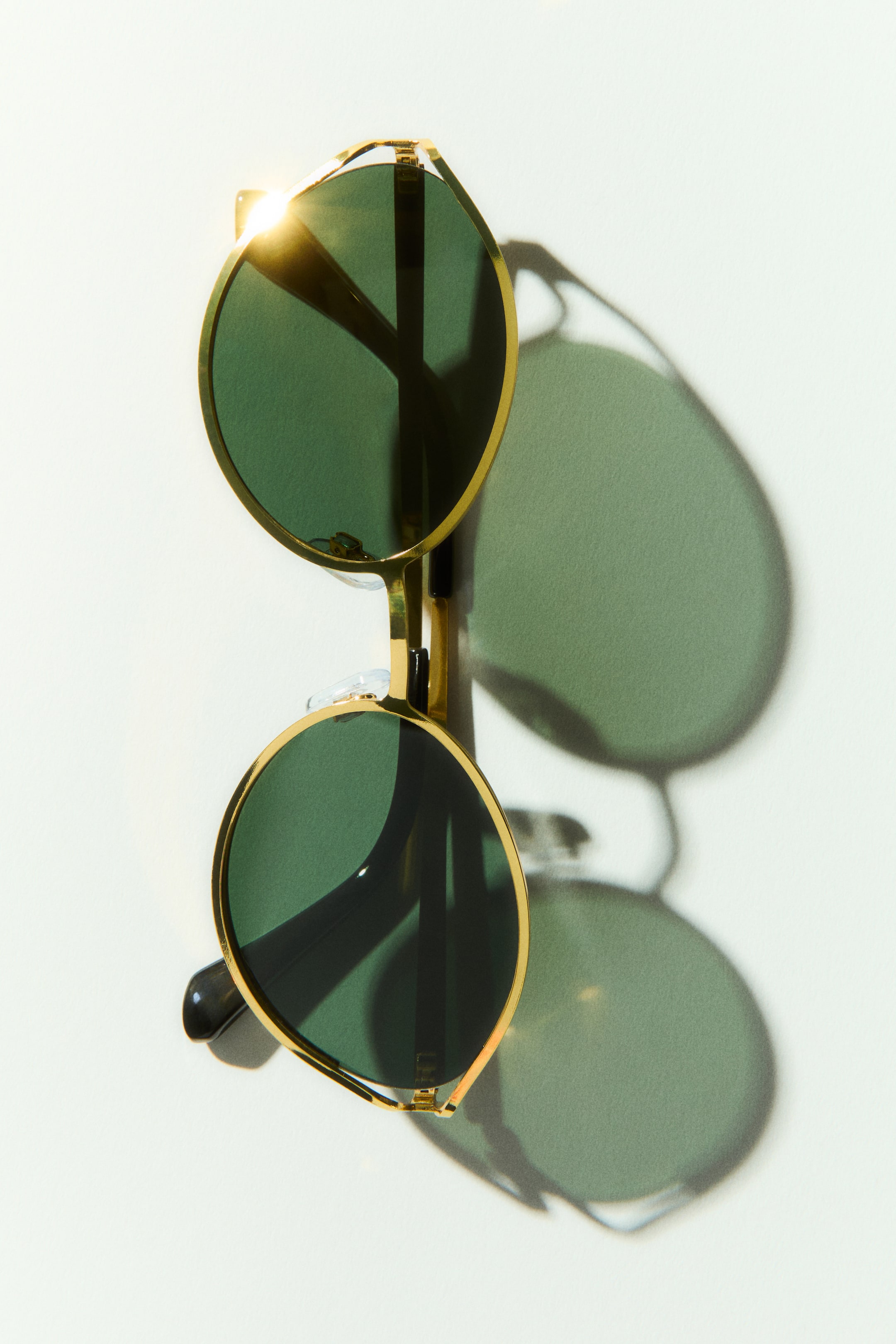 View larger image: Sunglasses feature round, dark green lenses set in gold-toned metal frames, with one lens reflecting bright light and clear nose pads visible on the bridge.