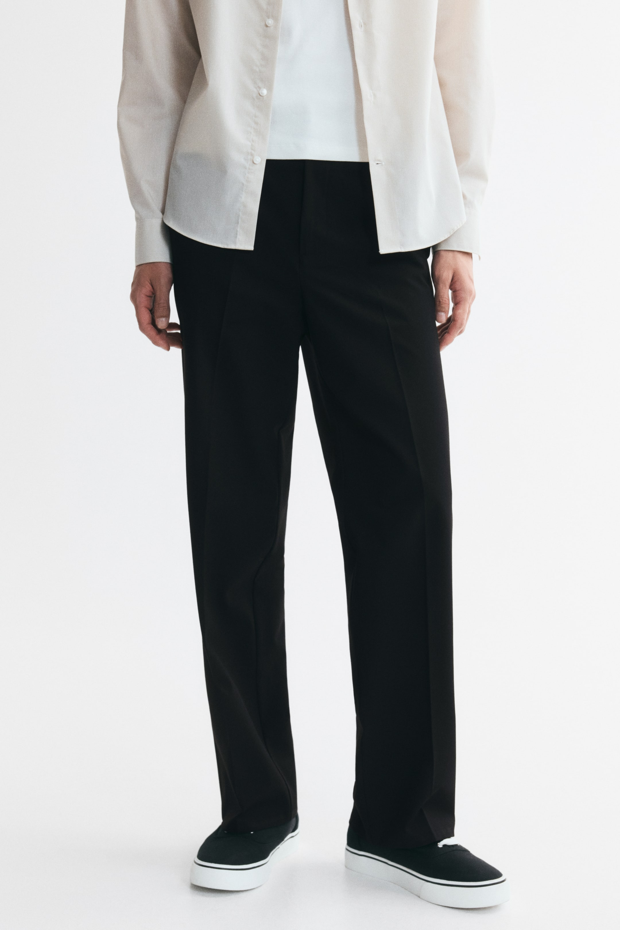 Prikaži veću sliku: Crne pantalone, sa naborima duž prednje strane nogavica i opuštenim krojem, nosi model. Model kombinuje pantalone sa svetlom košuljom i crnim patikama.