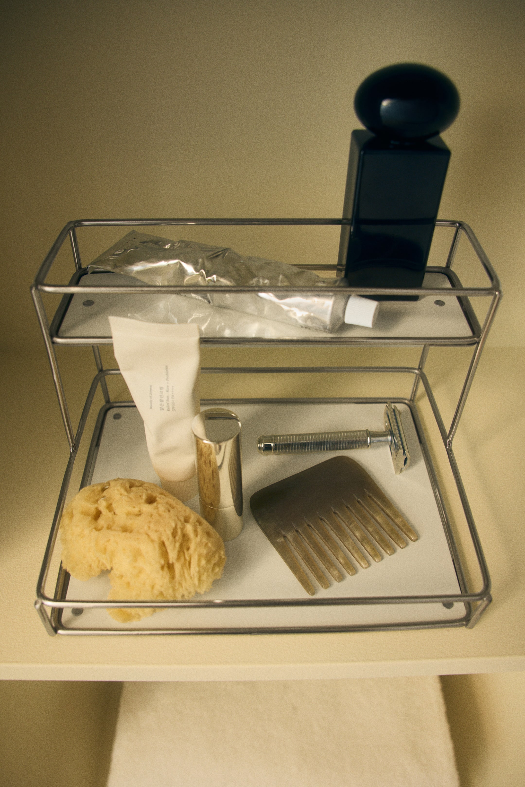 View larger image: A two-tier metal organizer is standing front-facing, featuring a silver wire frame with two flat white shelves. The top shelf holds a squeezed silver tube and a black bottle, while the wider bottom shelf holds a cream tube, a gold cylinder, a natural sponge, a wide-tooth comb, and a metal safety razor.