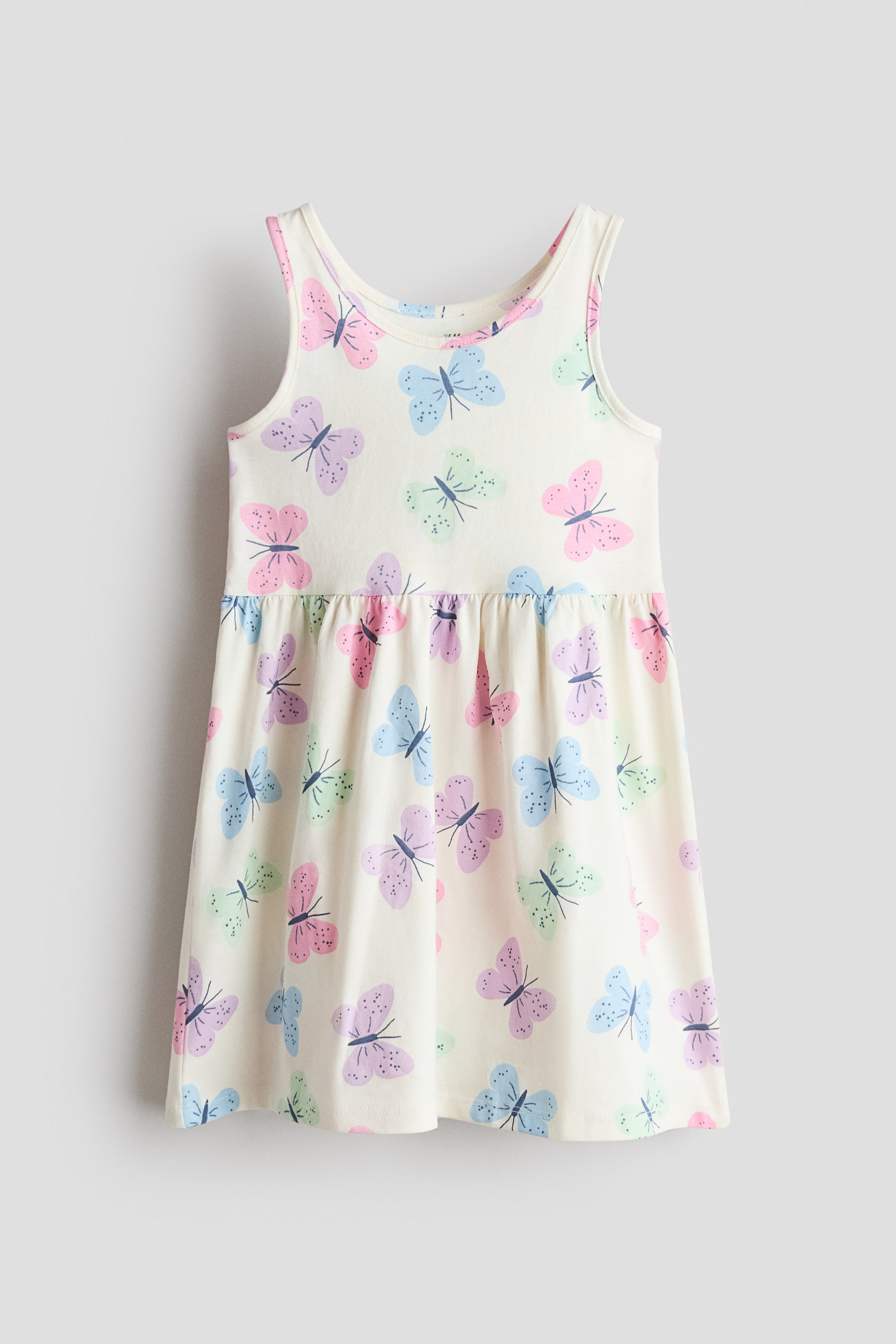 View larger image: The sleeveless dress is shown from the front, crafted from a light cream knit fabric adorned with pink, purple, blue, and green butterflies. It features a round neckline, a gathered seam at the waist, and a gently flared skirt.