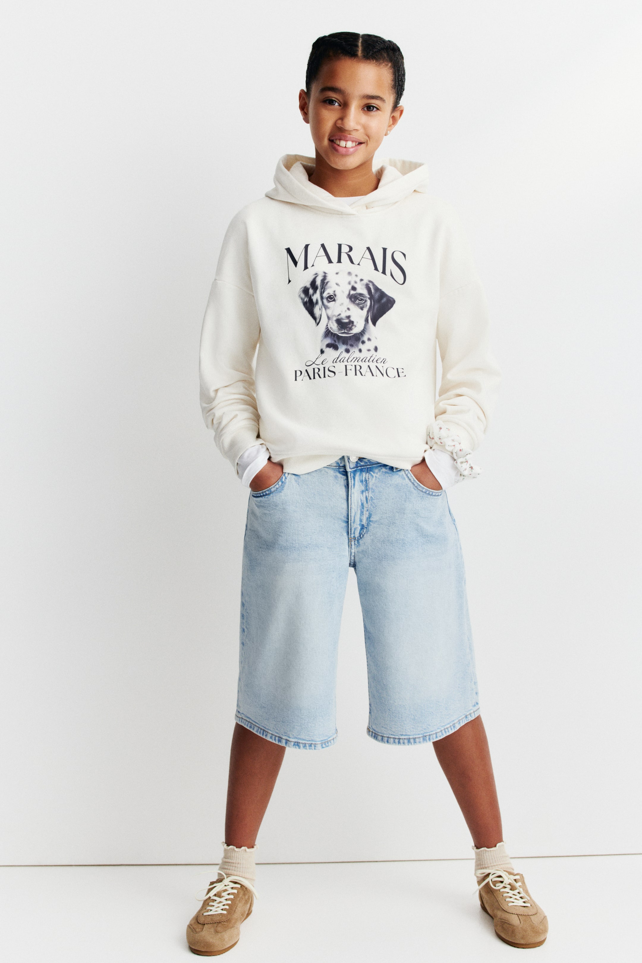 View larger image: A light beige hoodie is showcased from the front, featuring a hood and long sleeves. A black graphic print of a Dalmatian dog's head, with the text "MARAIS" and "PARIS FRANCE," is displayed on the chest. A white long-sleeve shirt is layered underneath, with its cuffs extending beyond the hoodie's cuffs.